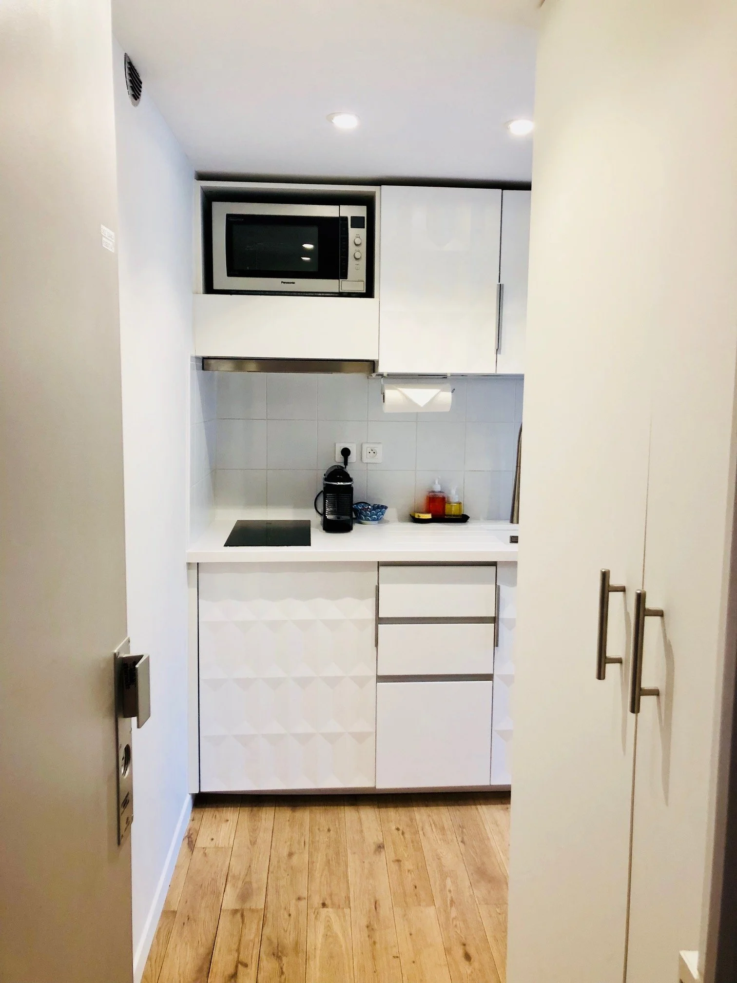 Small modern kitchen with white cabinetry, a microwave oven, a coffee machine, and a paper towel holder, with wood flooring and minimal decor.