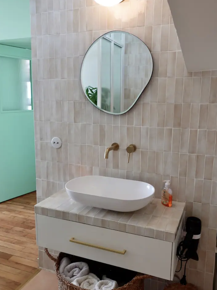 Bathroom sink with a white vessel basin on a tiled countertop, a circular mirror above, soap dispenser to the right, and a black hairdryer mounted on the wall. Under the sink is a basket with rolled towels. The wall has light-colored square tiles, an
