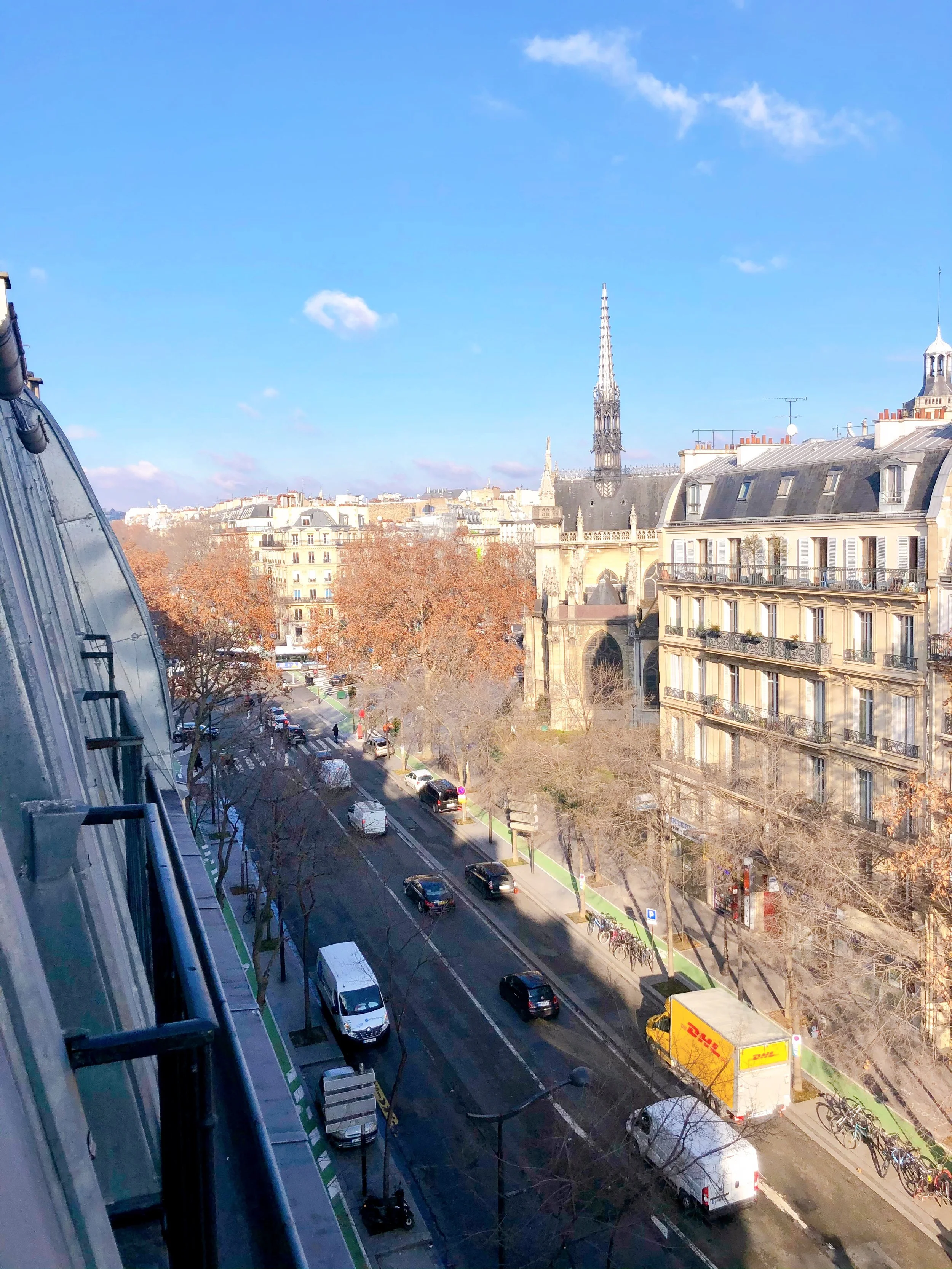 Paris city view with a street, cars, trees, old buildings with balconies, a church with a tall spire, under a blue sky with some clouds.