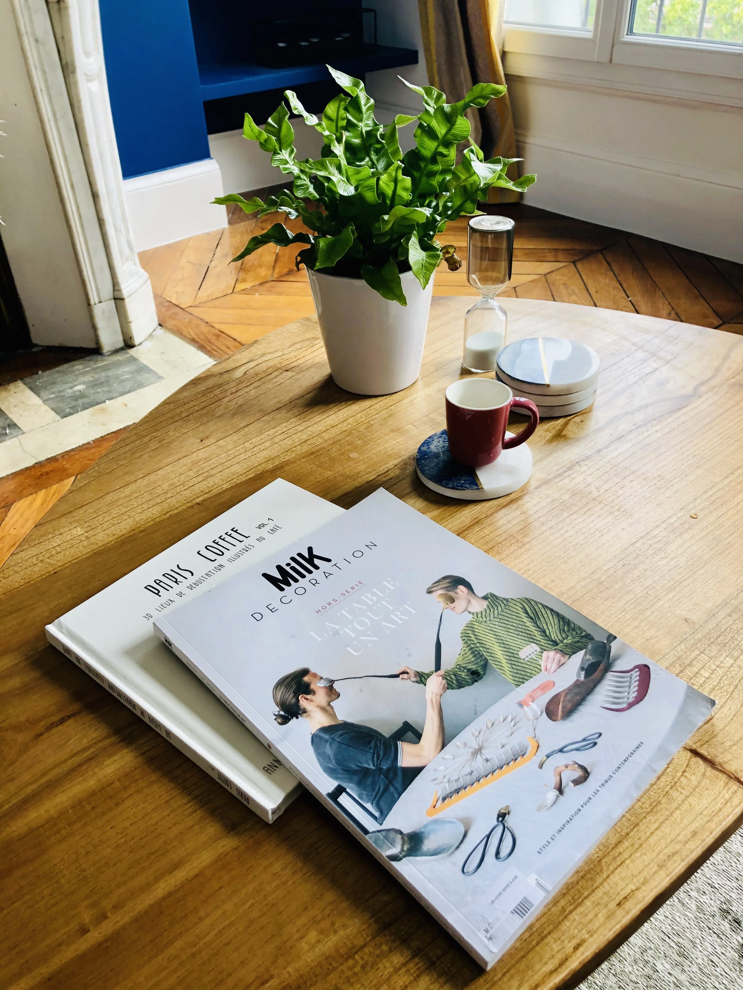 A wooden table with a magazine featuring a woman and a girl interacting, a red coffee mug on a coaster, an hourglass, a small container, and a white dish. A potted green plant, a window with sunlight, and a blue bookshelf in the background.