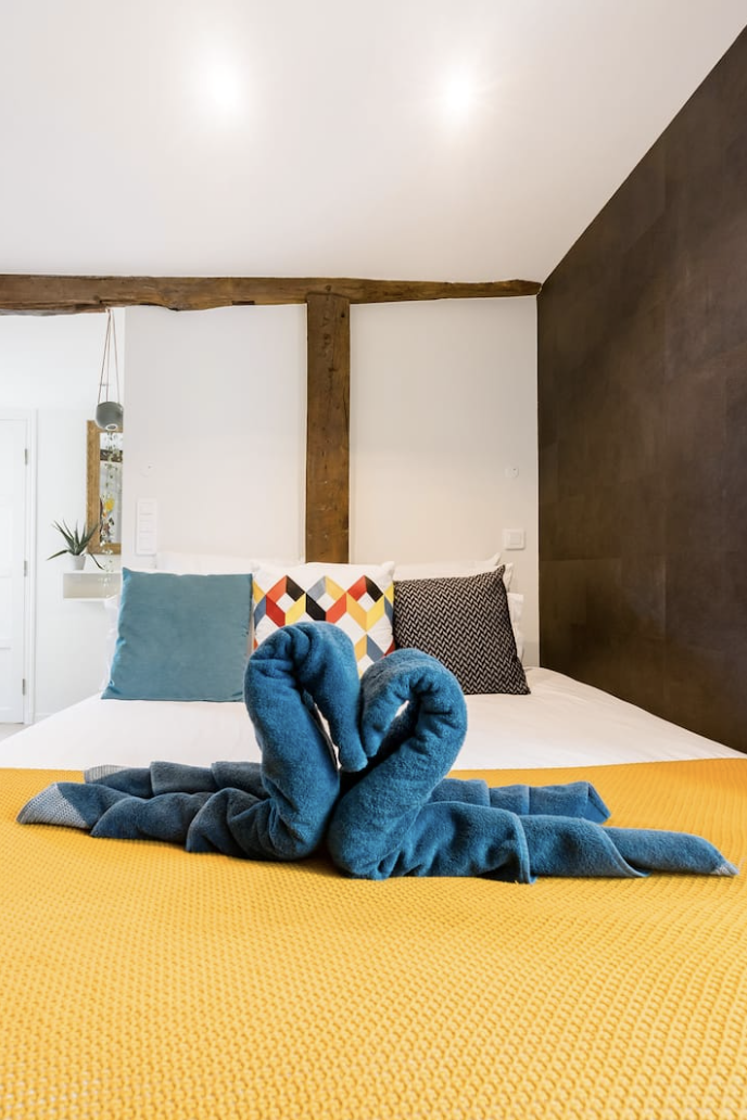 A neatly made bed with colorful pillows, including a geometric-patterned pillow, a teal pillow, and a black and white patterned pillow. A towel art sculpture resembling a swan is placed on a yellow blanket at the foot of the bed. The room has a woode