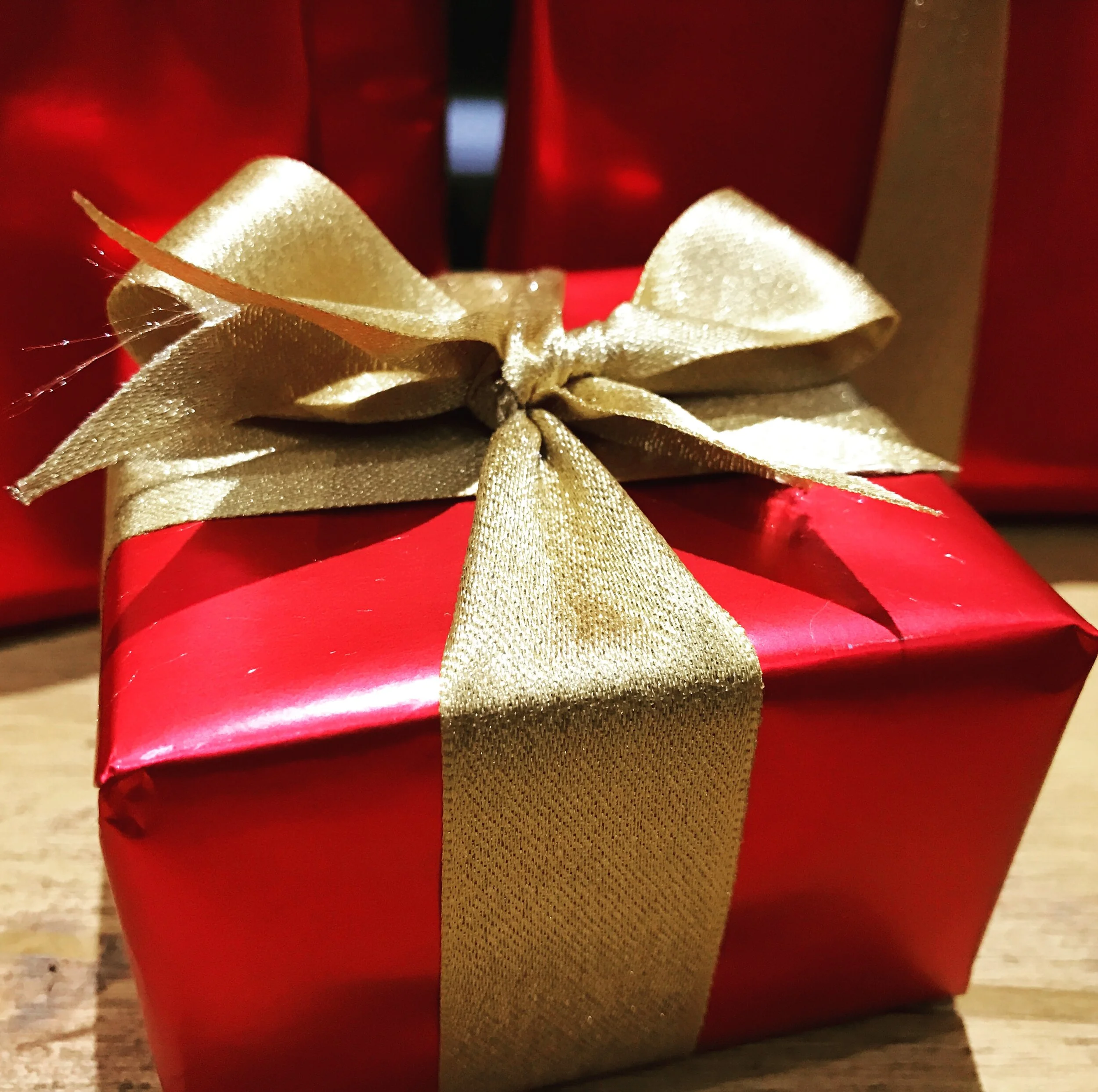 A red gift box wrapped with a gold satin ribbon tied in a bow, sitting on a wooden surface with other red gift boxes in the background.