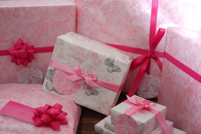 Multiple pink and white gift boxes wrapped with pink ribbons and bows, surrounded by pink wrapping paper decorated with floral and butterfly patterns.