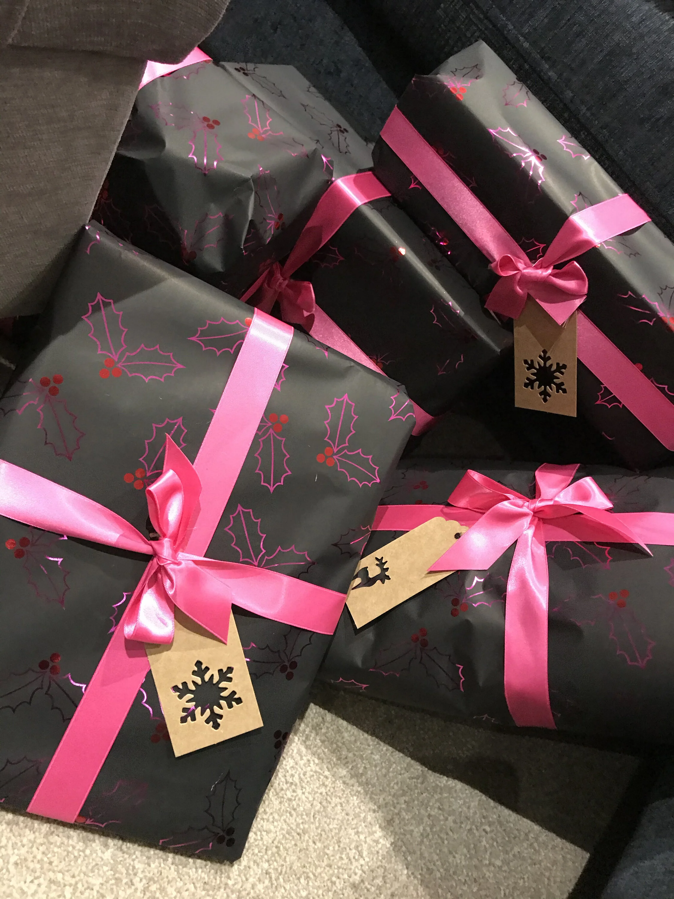 Several Christmas presents wrapped in dark paper with holly and berry designs, tied with bright pink ribbons and pink bows, with holiday-themed tags attached.