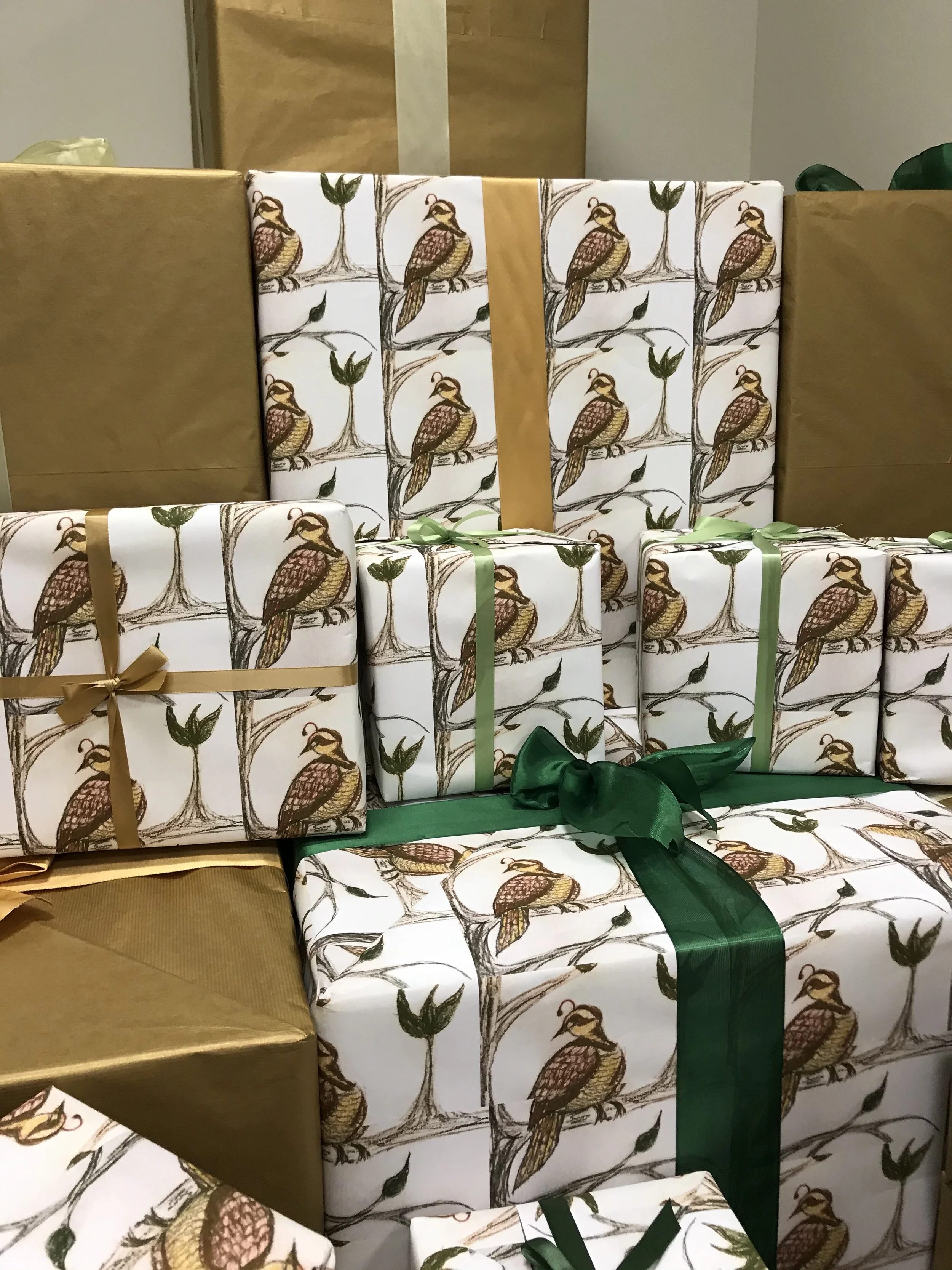 Stacked Christmas presents, some wrapped in white paper with a bird and tree pattern, others in gold paper, some with green or gold ribbons, on a green surface.