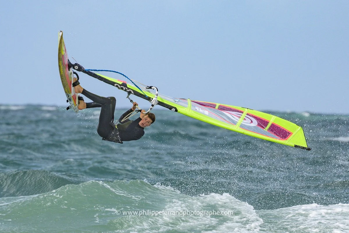 Un windsurfer rookie effectuant une manœuvre acrobatique au-dessus de la mer, portant une combinaison de plongée noire, avec une voile colorée jaune, rose et blanche.