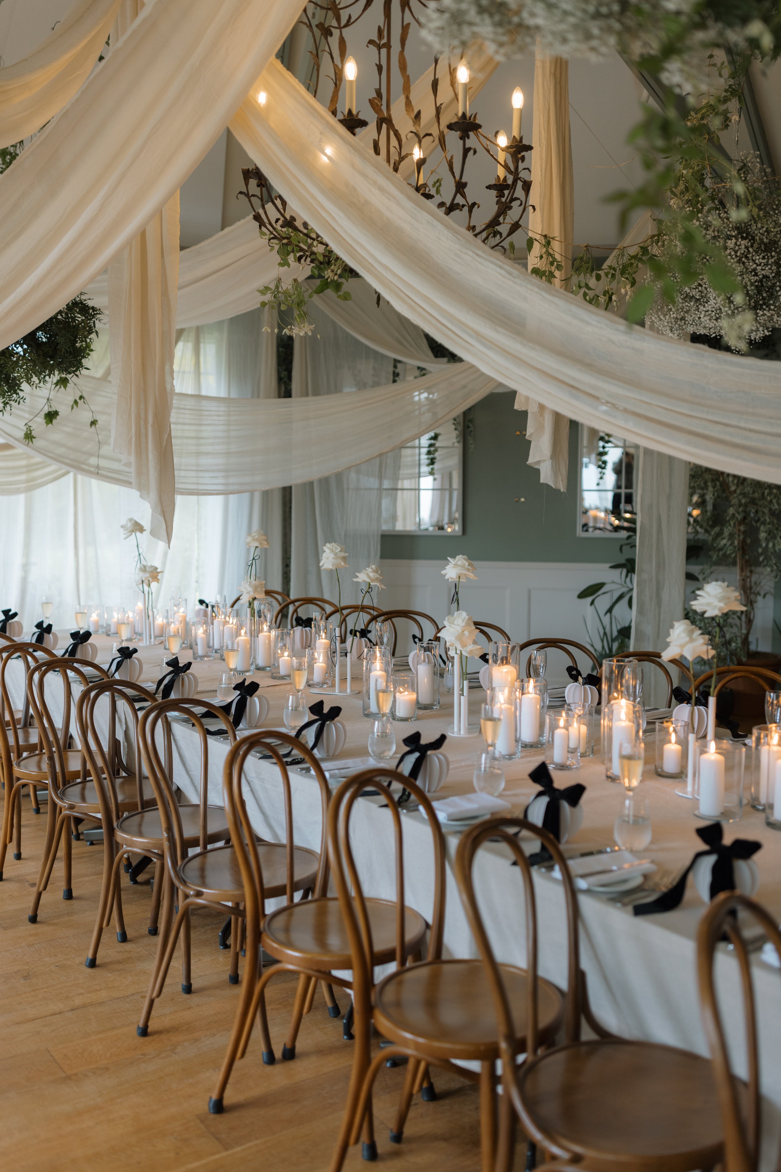 A decorated event space with a long dining table set for guests, featuring candles, white flowers, and black ribbon accents. The ceiling is adorned with elegant draped white fabric and a chandelier, with greenery and floral arrangements hanging from above.