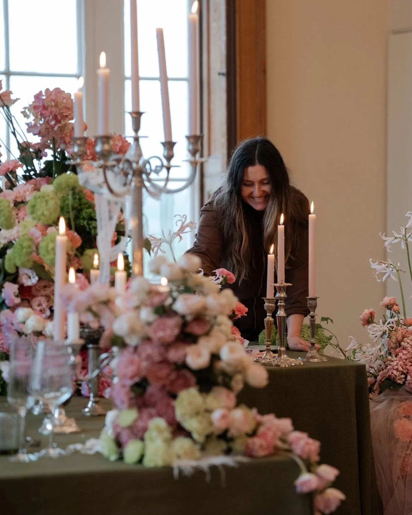 Woman setting up a table decorated with pink and white flowers and tall candelabras with lit candles in a warmly lit room.