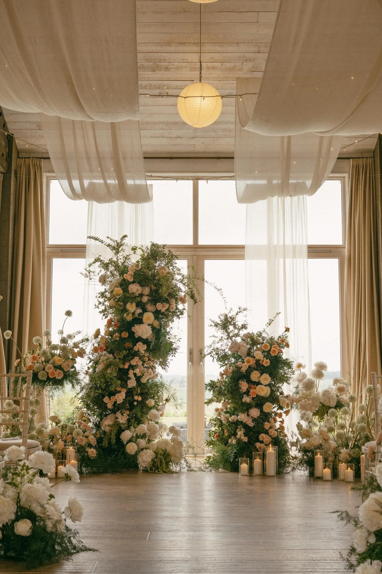Elegant wedding altar decorated with tall floral arrangements and candles, located indoors in front of large windows with sheer curtains and draped fabric ceiling.