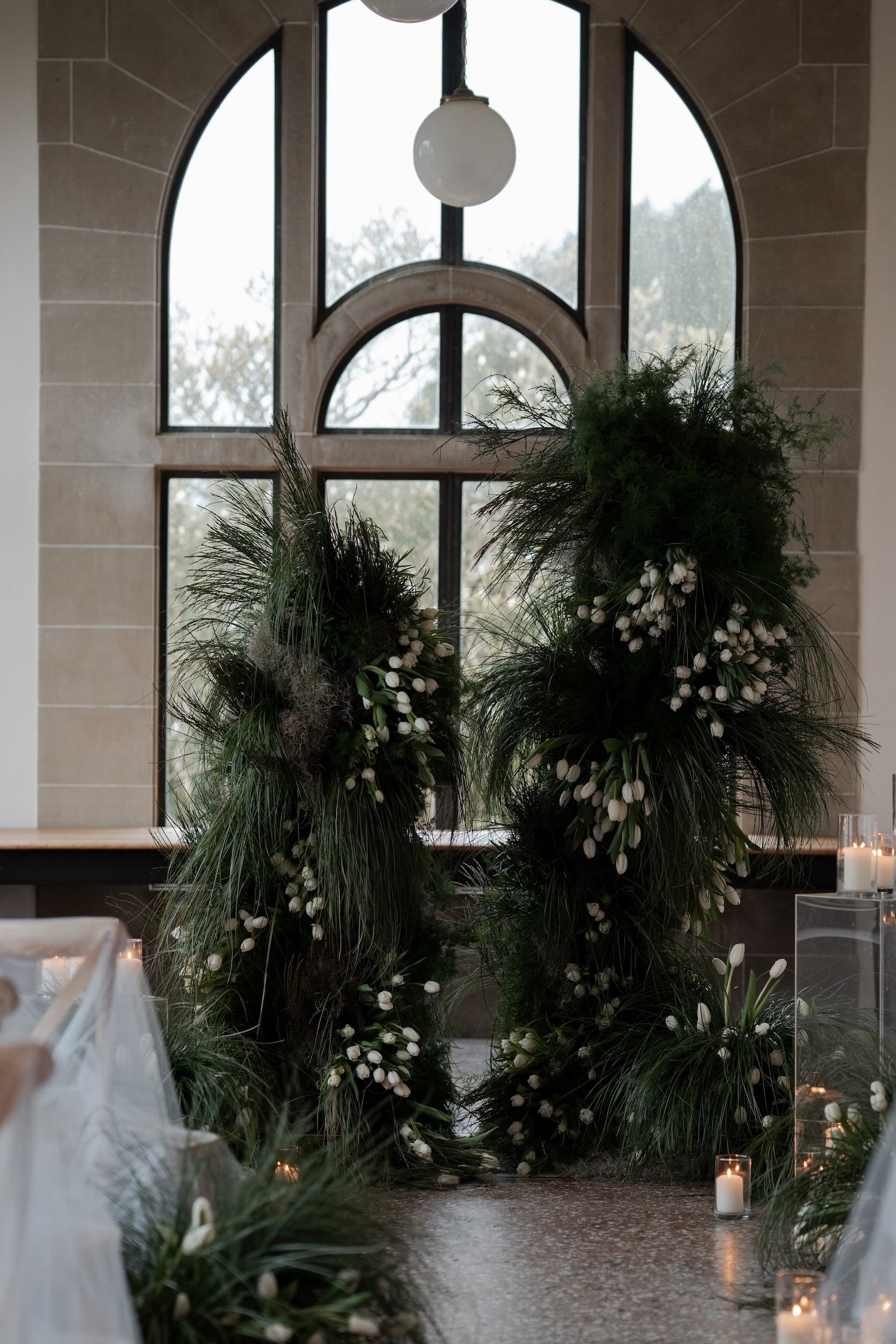 Indoor scene with tall, lush greenery arrangements with white flowers, candles, and a decorative window with arched panes in the background.