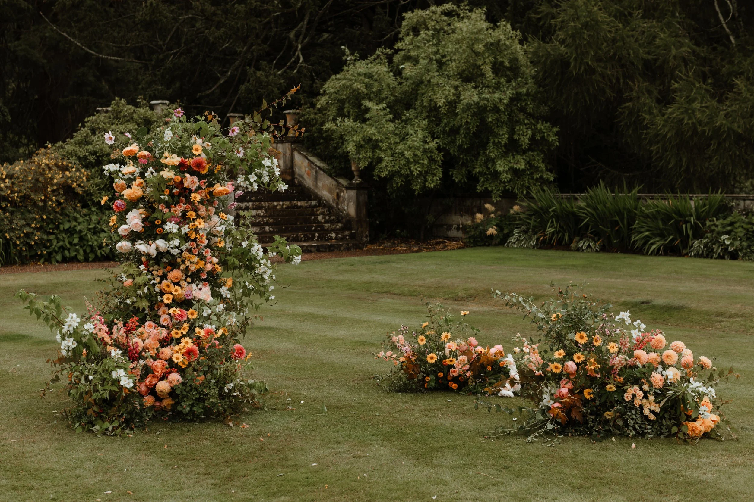 A garden scene featuring a large floral arrangement with pink, white, orange, and yellow flowers on grass, with stone stairs and lush green trees in the background.
