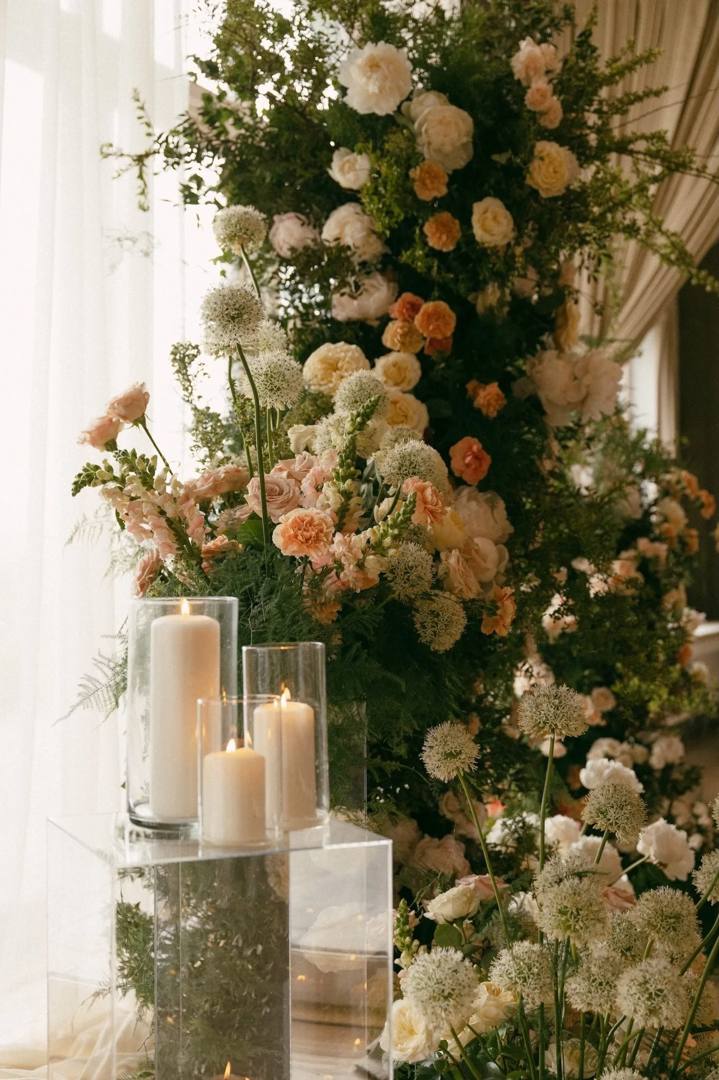 A large floral arrangement with white and peach-colored flowers, greenery, and some spherical white blooms. In front of the arrangement are three white candles inside glass holders on a clear acrylic stand, with some candles lit.