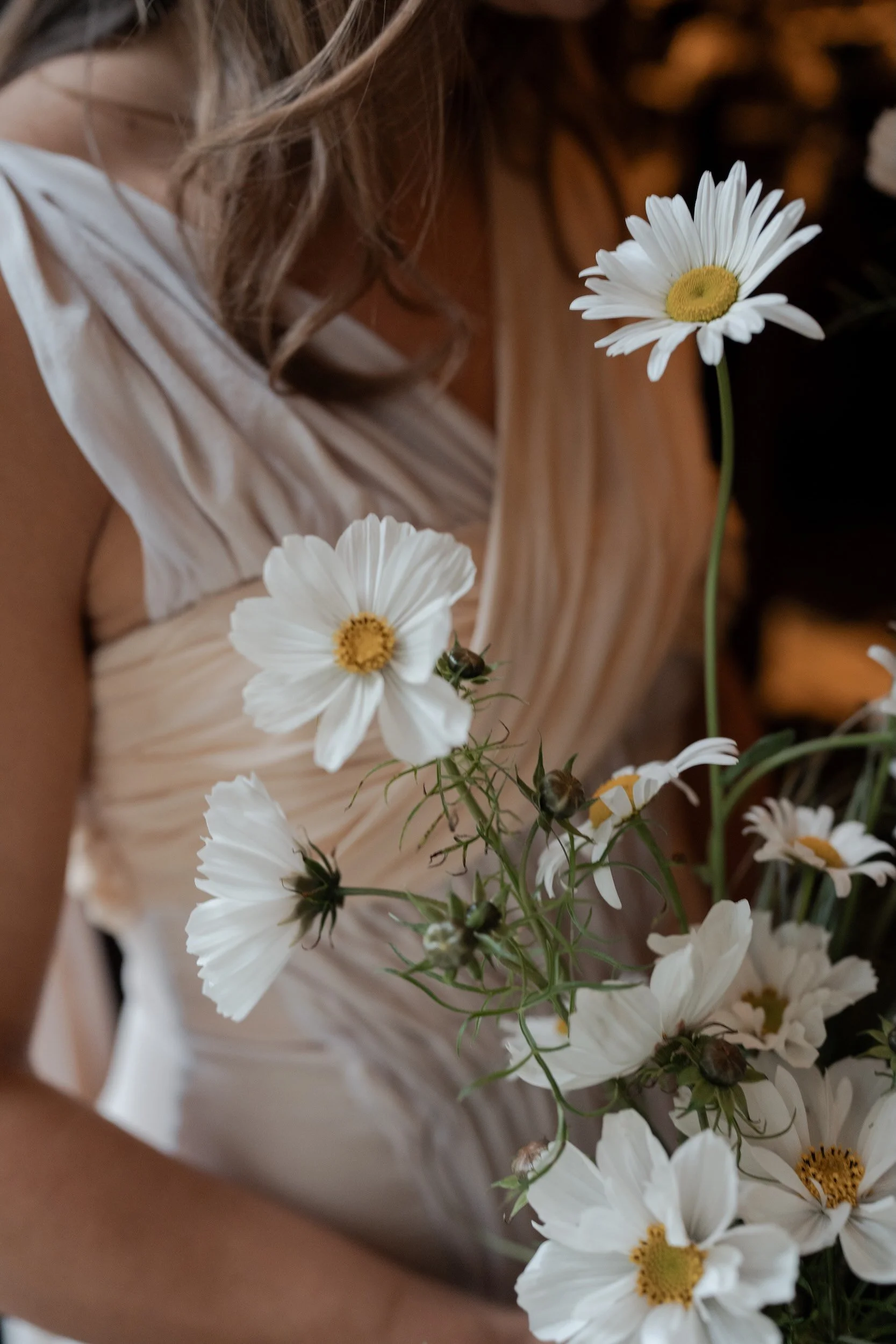 Close-up of a woman holding a bouquet of white daisies with yellow centers, wearing a light-colored, pleated dress.