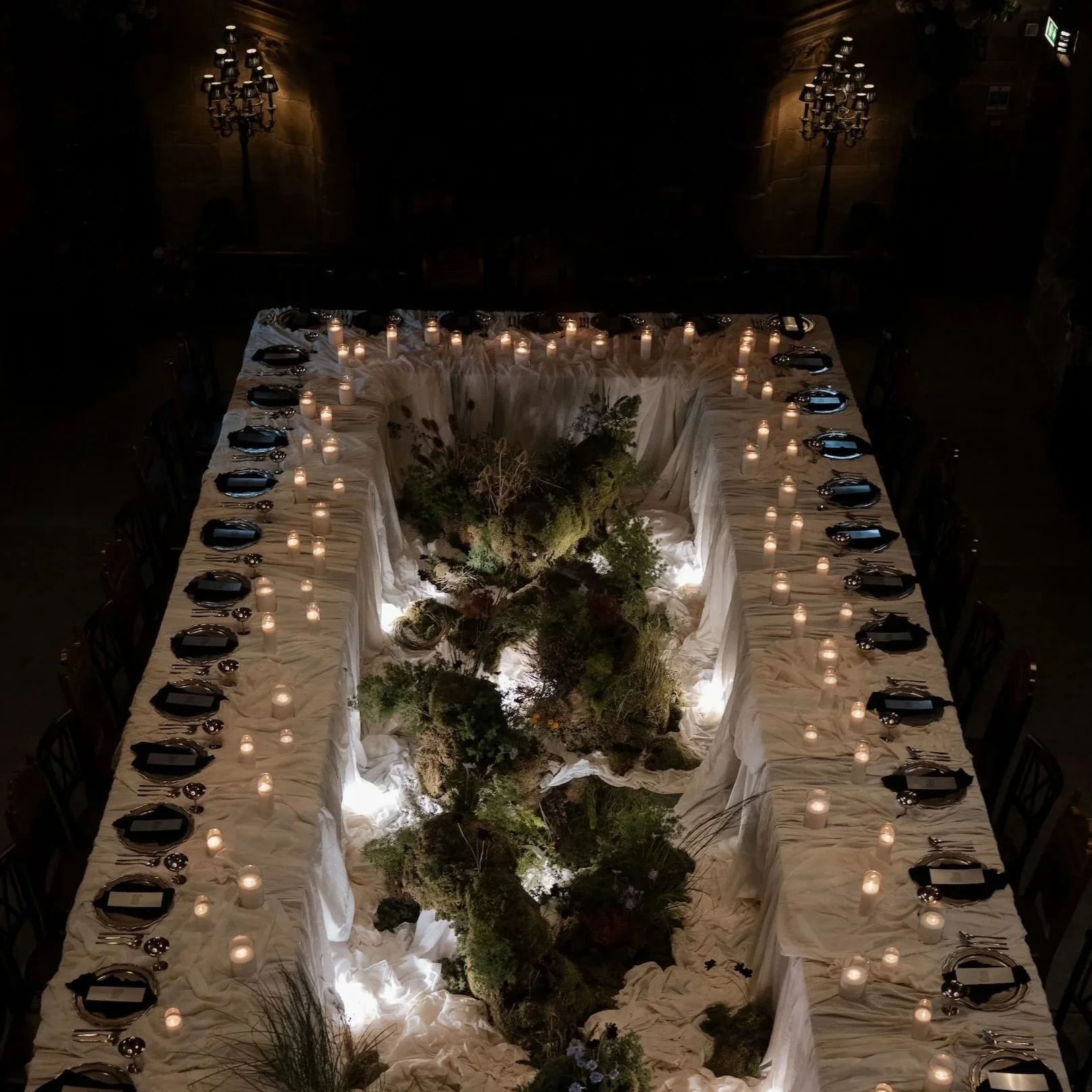 A long, rectangular dining table decorated with white tablecloths, surrounded by chairs, with numerous lit candles and greenery running down the center, in a dimly lit room with ornate wall sconces.