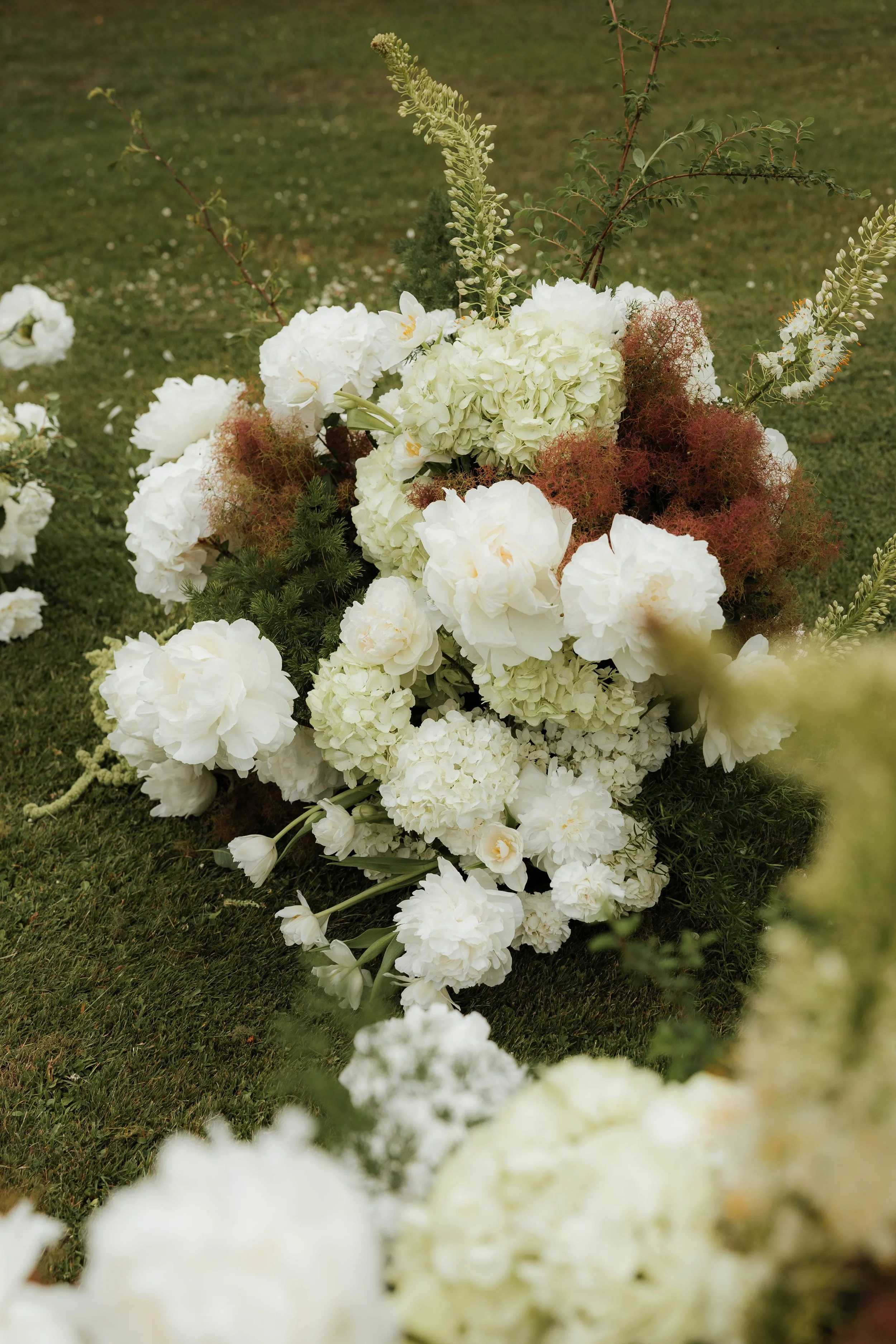 White floral arrangement with hydrangeas, peonies, and greenery on grass.