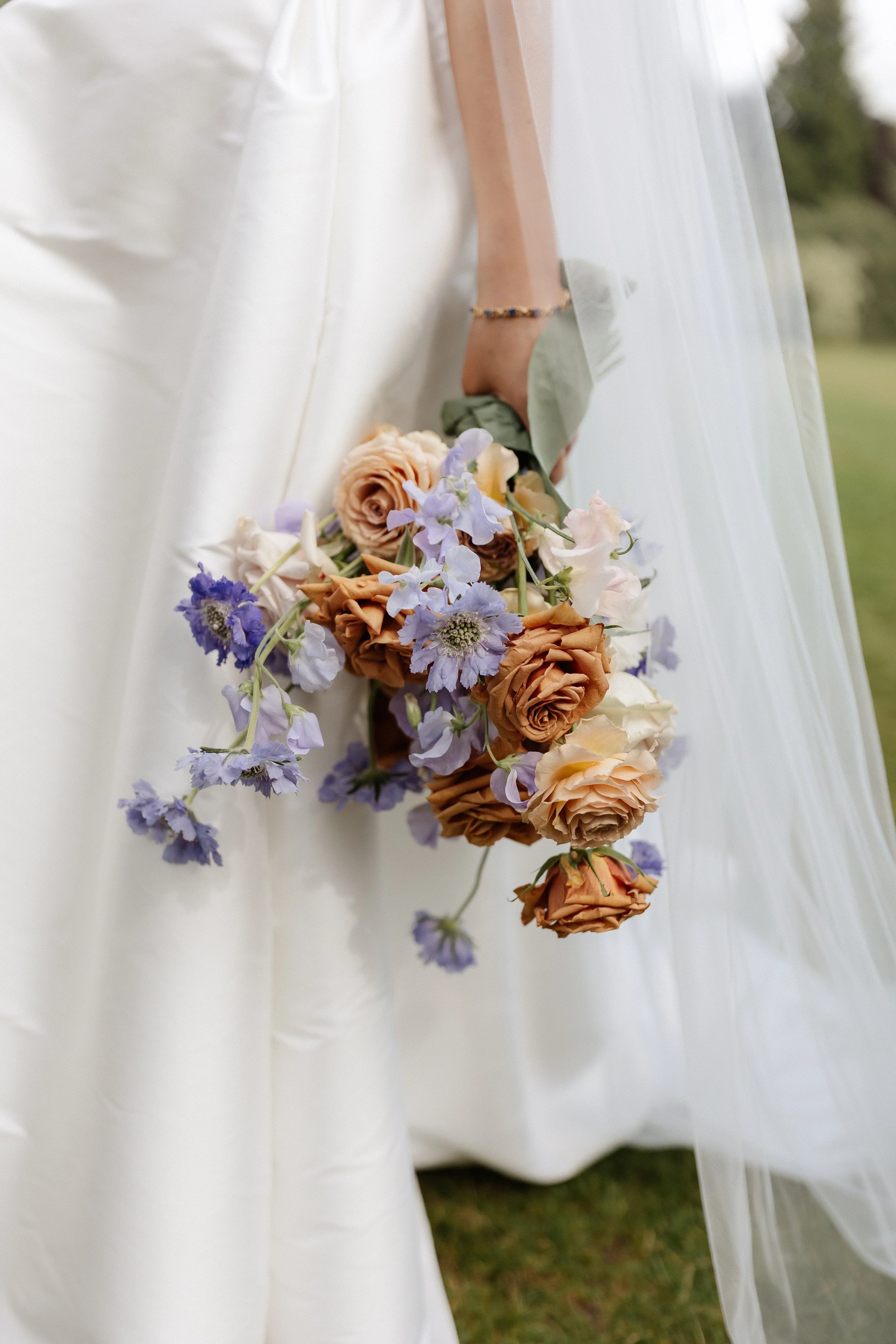 Close-up of a bride holding a cascading bouquet of peach, cream, and lavender flowers, with a white wedding dress and veil in the background.