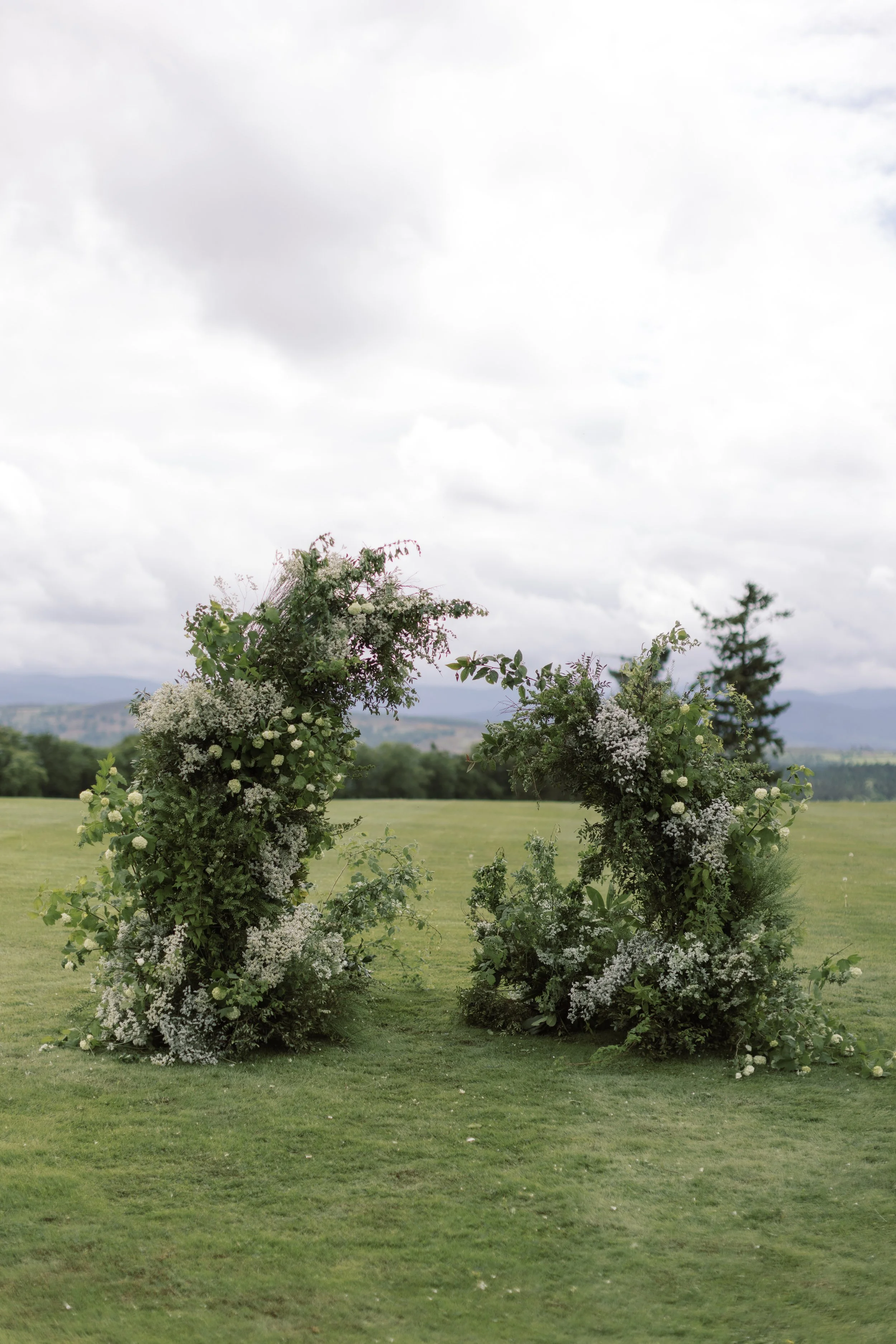 Two large floral arrangements made of white and green flowers standing outdoors on a grassy field with a cloudy sky overhead.