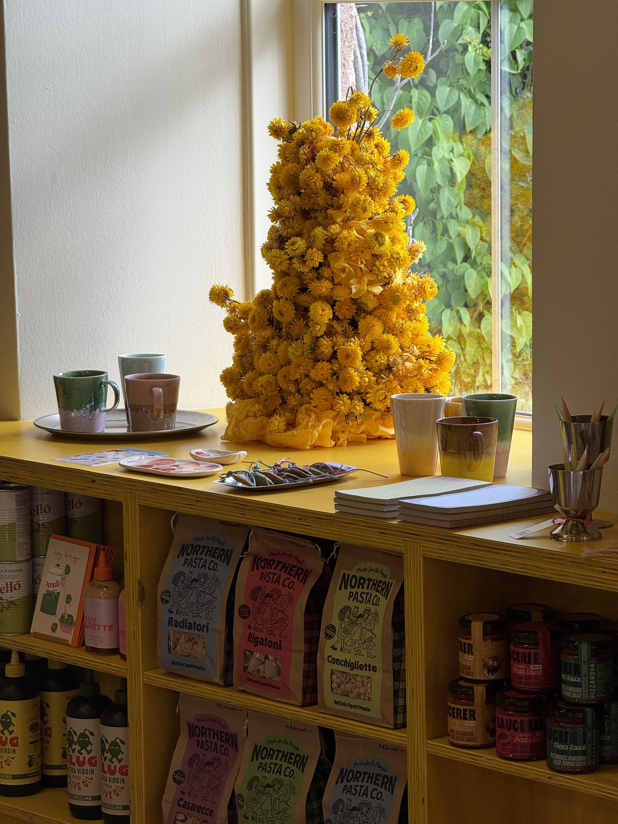 A yellow table with a yellow shelving unit, featuring a large yellow floral centerpiece on the windowsill, with tea cups on a tray, various packets of pasta, and spice jars below and around.