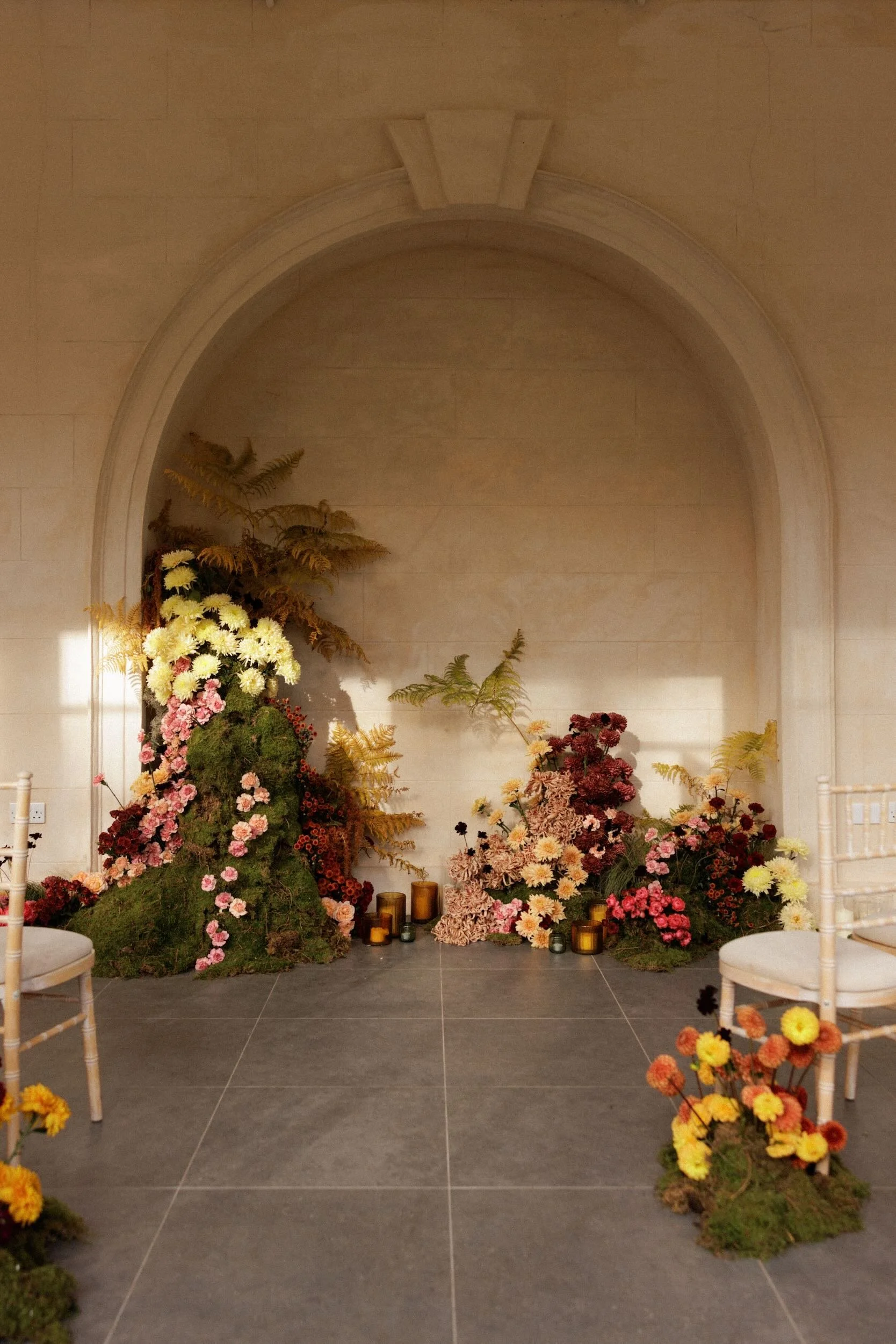 Indoor floral display with various flowers and candles against a beige wall and arched niche.