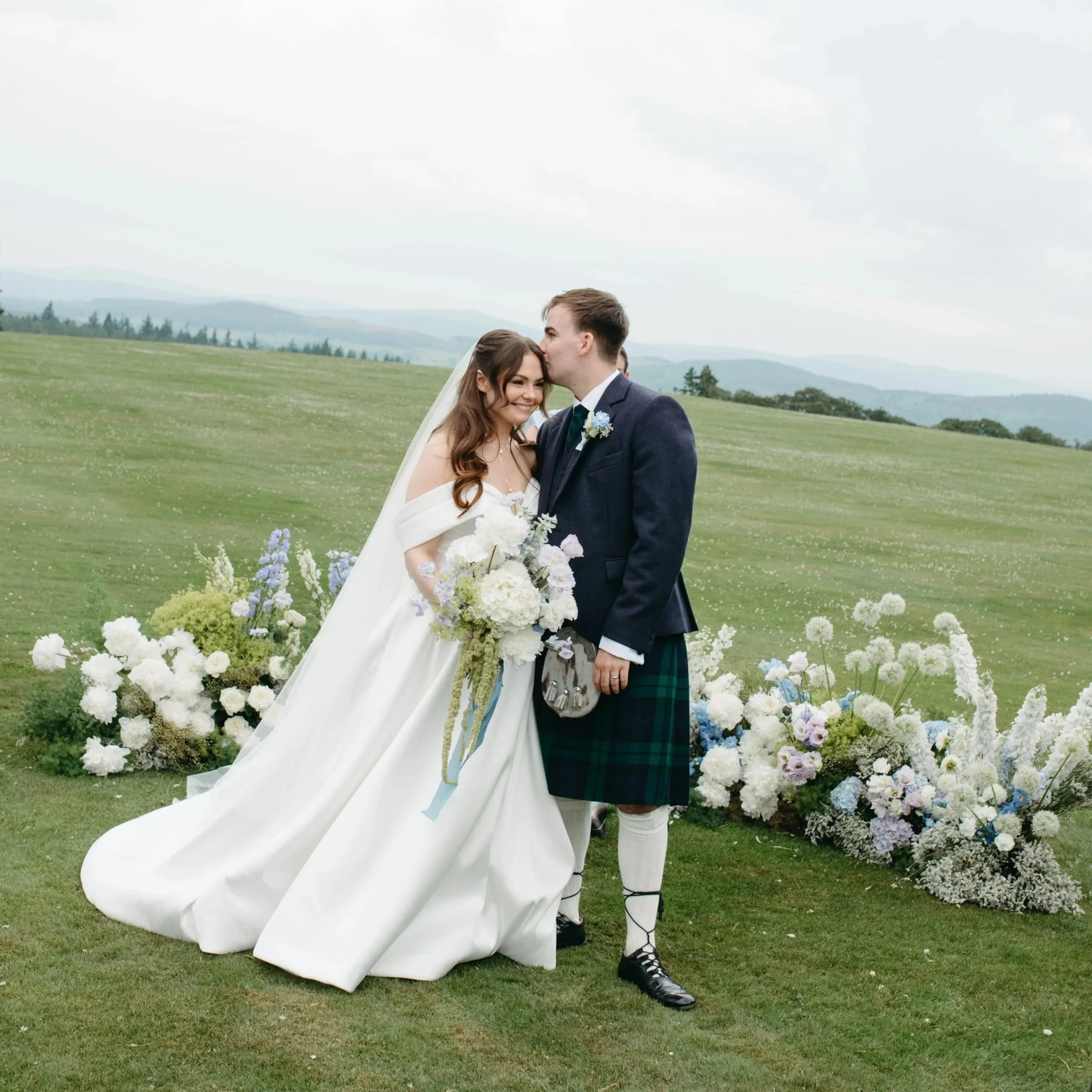 A bride and groom share a kiss outdoors during their wedding ceremony. The bride is holding a bouquet of white and purple flowers, and the groom is wearing a traditional Scottish kilt and jacket. They are standing in front of a floral arch made of white, purple, and green flowers on a grassy field with a mountain landscape in the background.