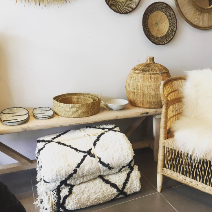 Décor intérieur avec paniers en osier, une table en bois, un tapis blanc avec des motifs noirs, un fauteuil en osier avec une couverture en fourrure.