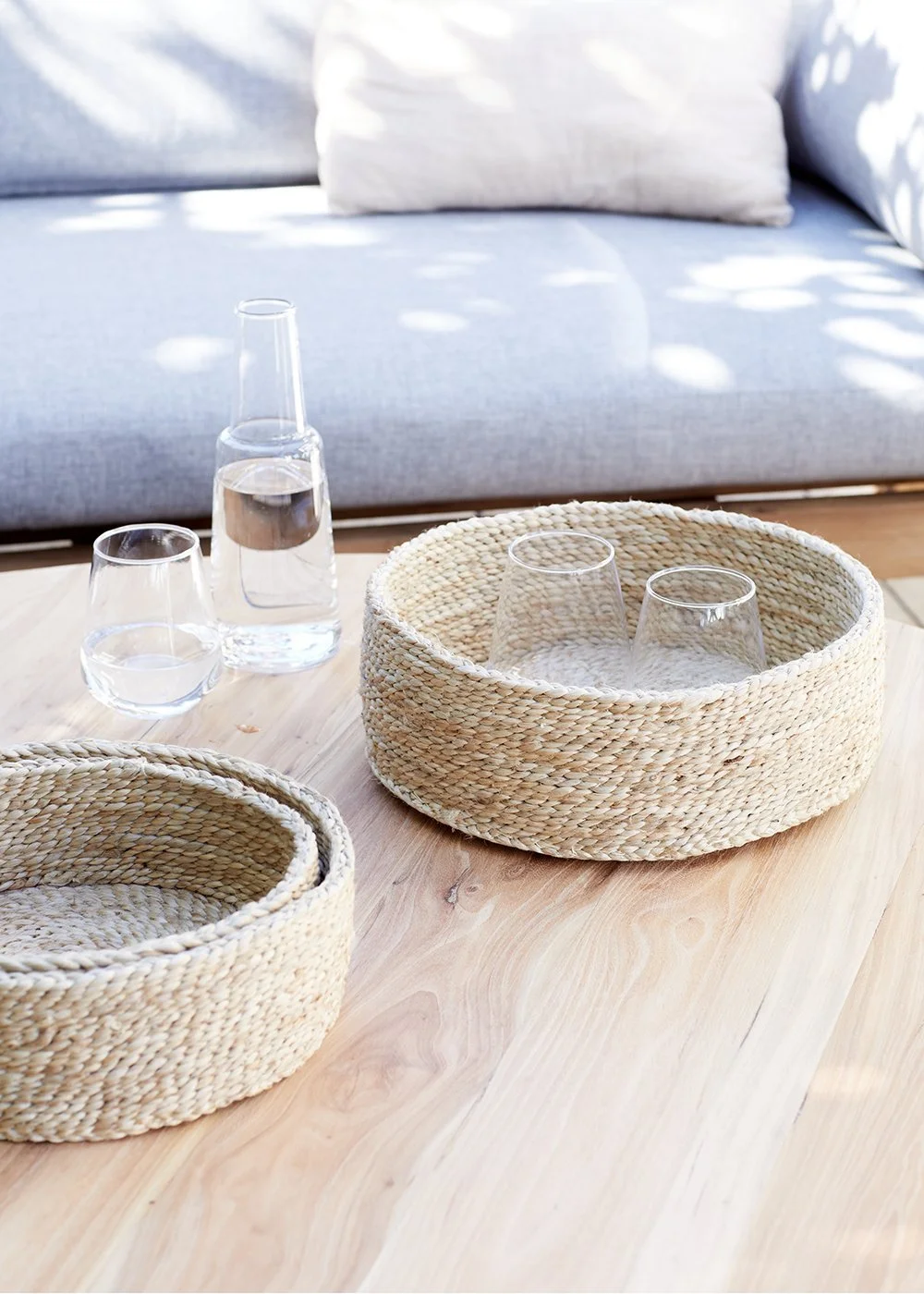 Table en bois clair avec deux paniers en osier, une carafe d'eau, deux verres d'eau et deux verres à vin vide, devant un canapé gris avec coussin blanc, dans un salon lumineux.