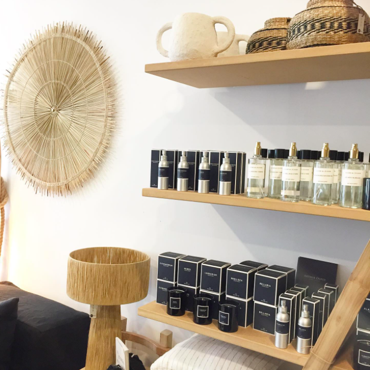 Étagère en bois avec des flacons de parfum, un pot blanc, des paniers tressés et un grand décor en fibre naturelle suspendu sur un mur blanc.