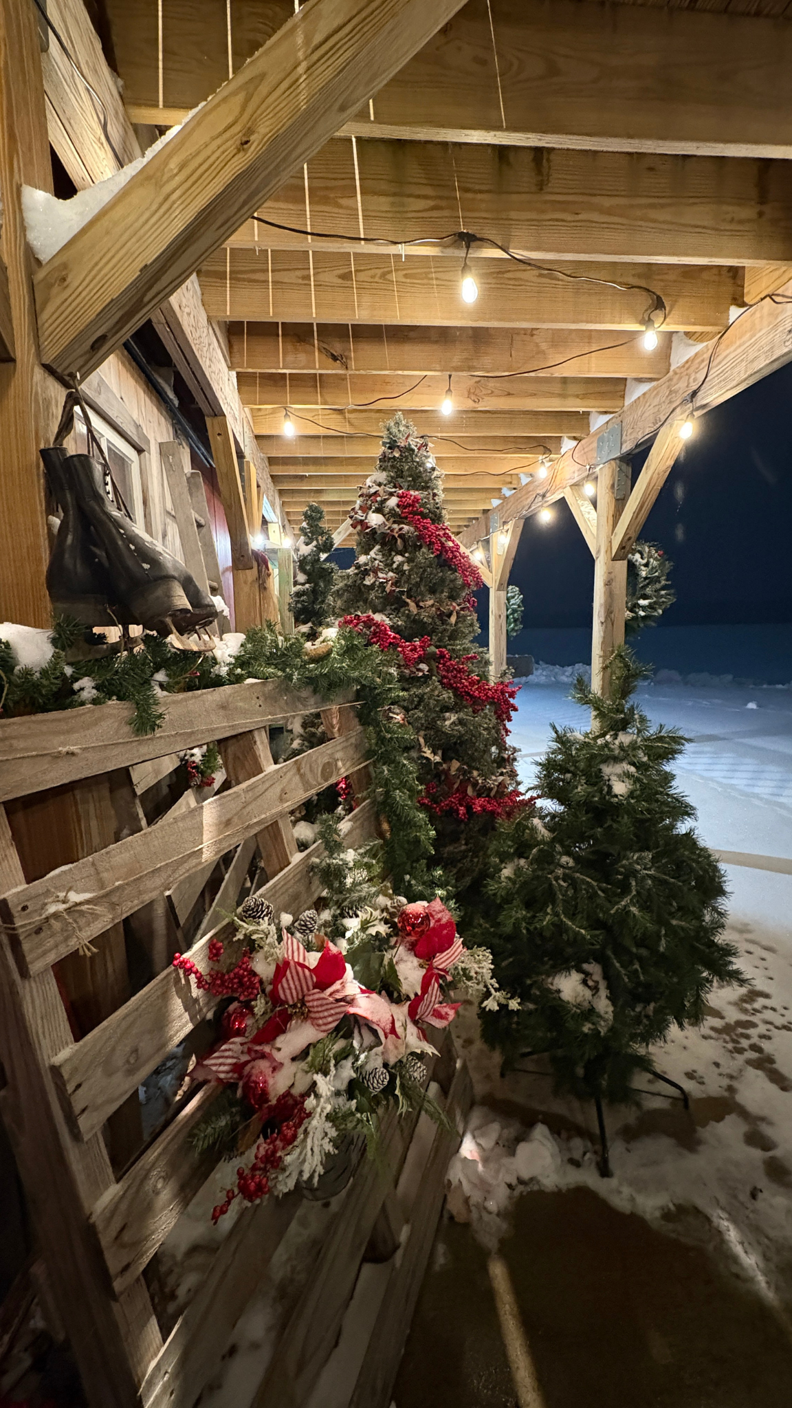 Decorated Christmas tree with red garland, Christmas flowers, and snow on the ground, on a covered outdoor porch at night with string lights.