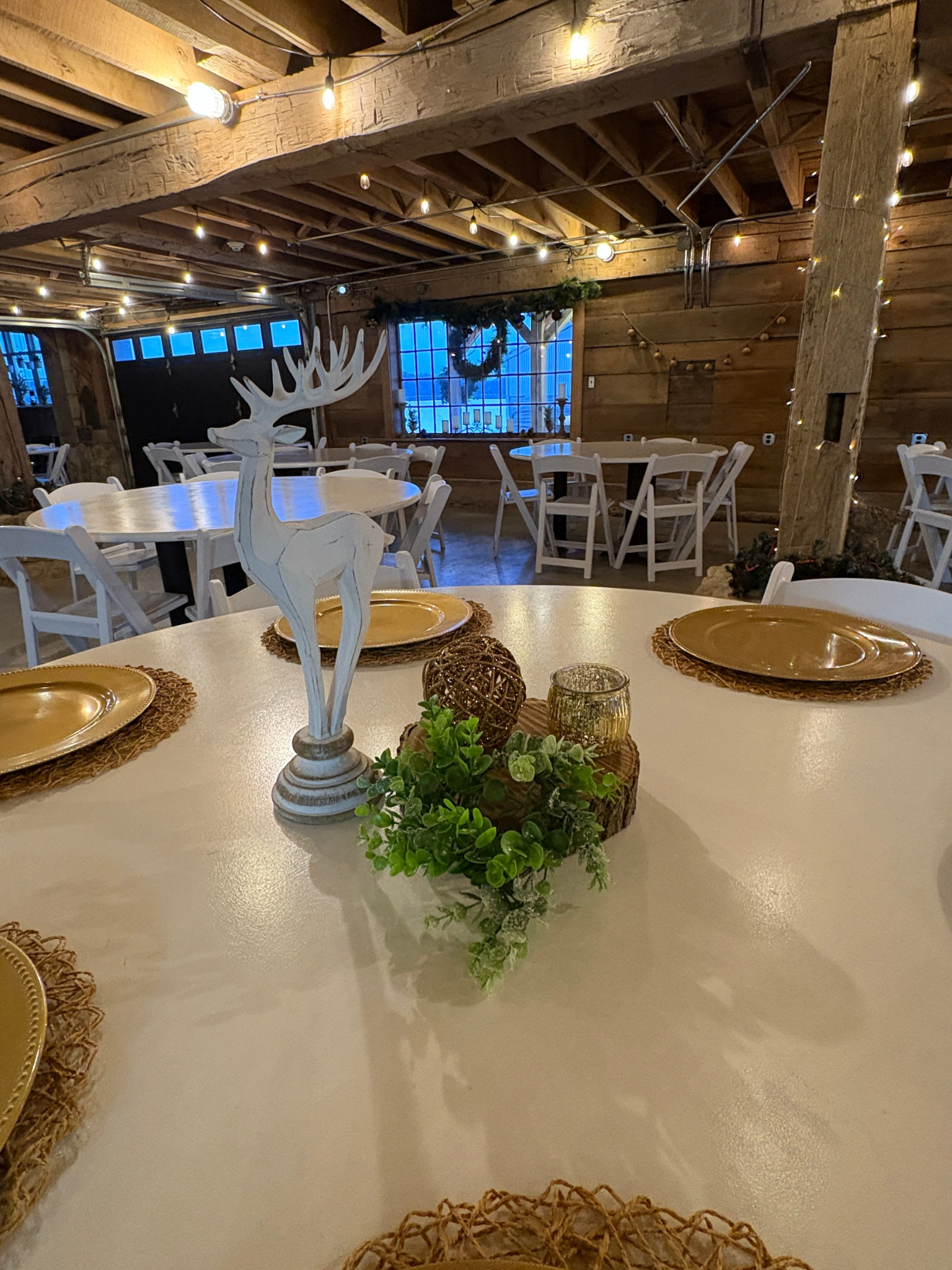A rustic indoor dining space decorated for Christmas with string lights, a wooden reindeer centerpiece, and greenery on the table, featuring wooden walls, large windows, and white chairs.