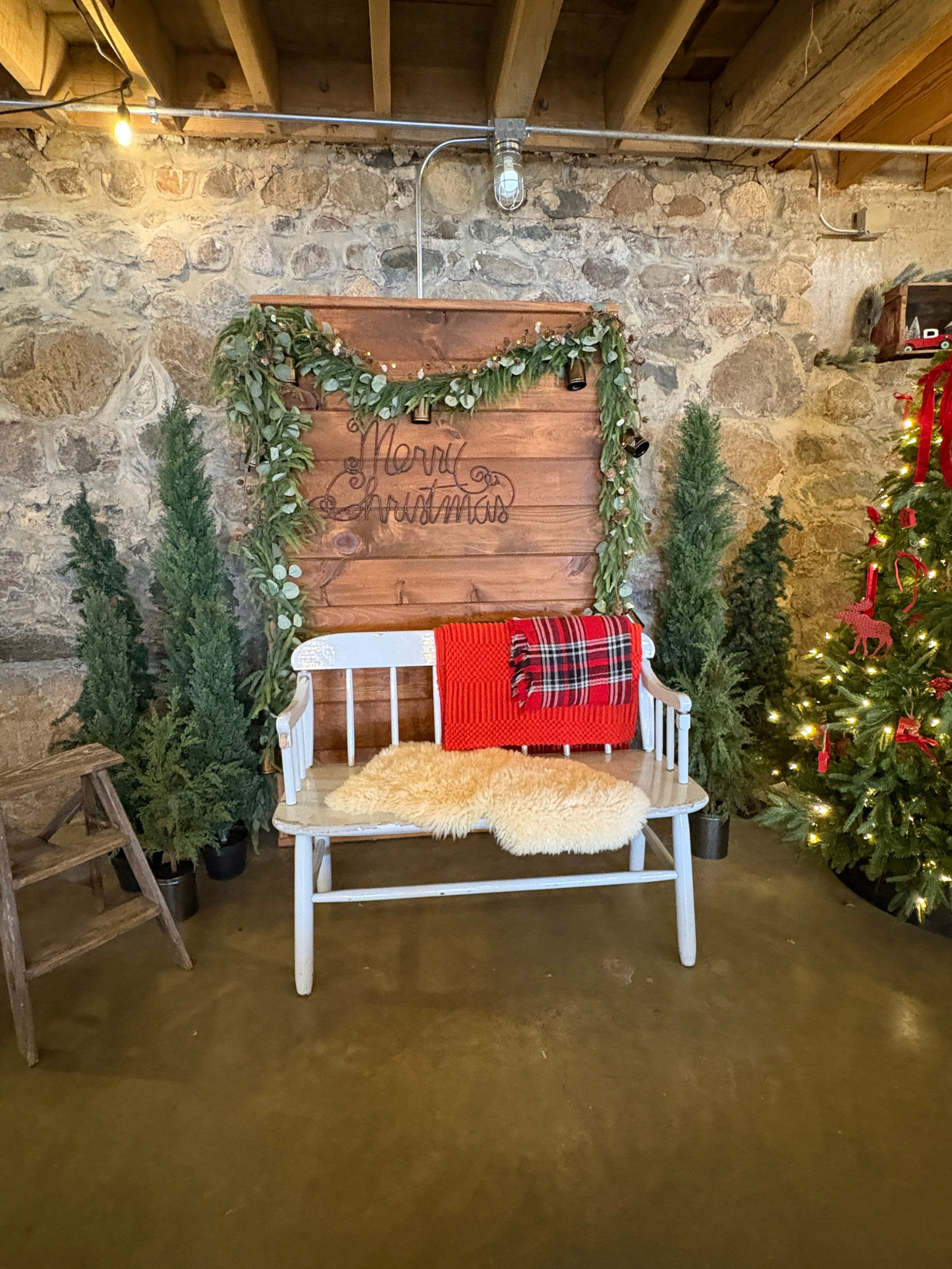 A rustic Christmas scene with a white wooden bench covered with a cream fur throw and a red plaid blanket, against a stone wall decorated with a wooden sign reading 'Merry Christmas' and garland, surrounded by small evergreen trees and a decorated Ch