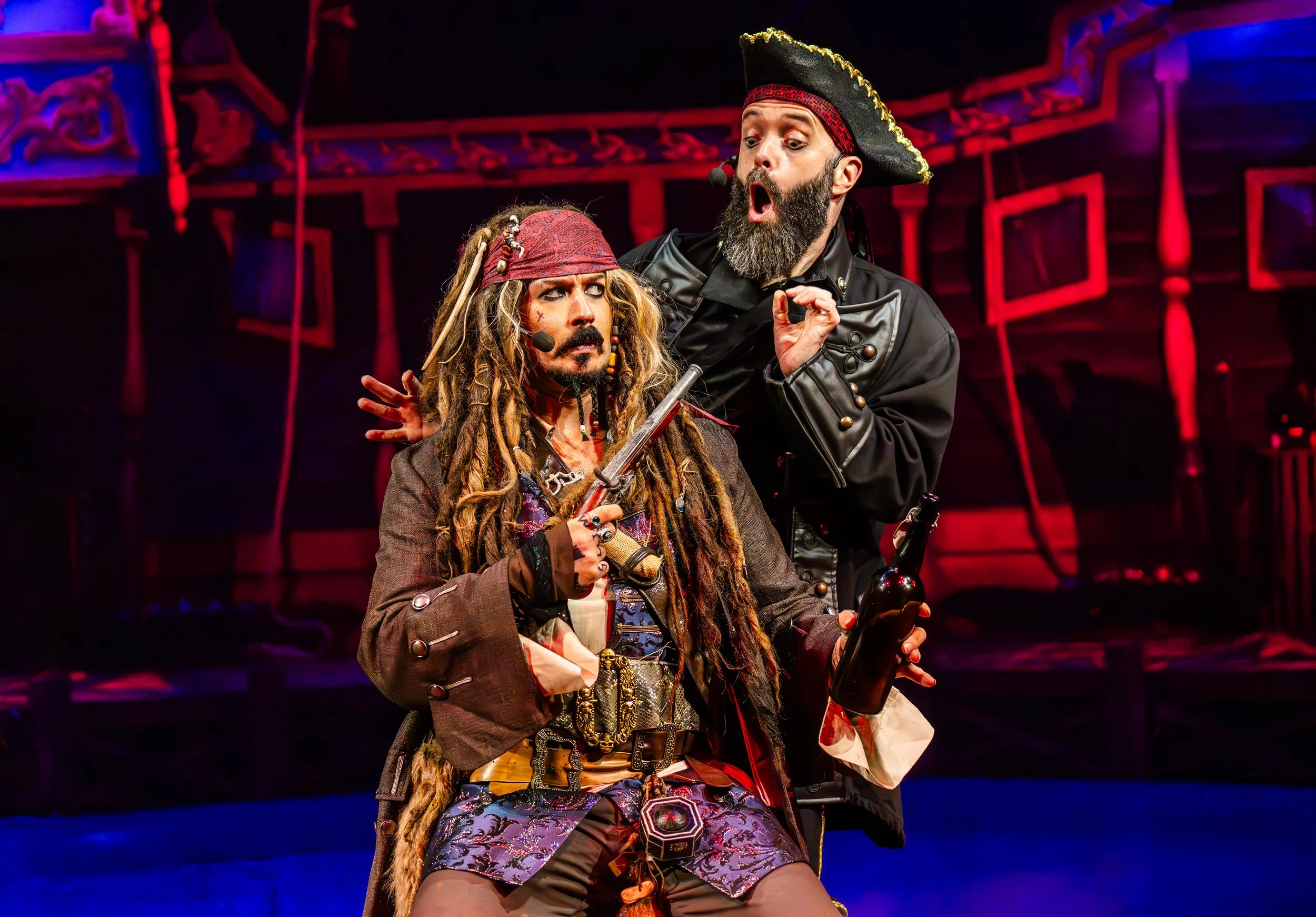 Two actors performing on stage, dressed as pirates with theatrical expressions, one holding a sword and the other a bottle, with a dramatic red and blue background.