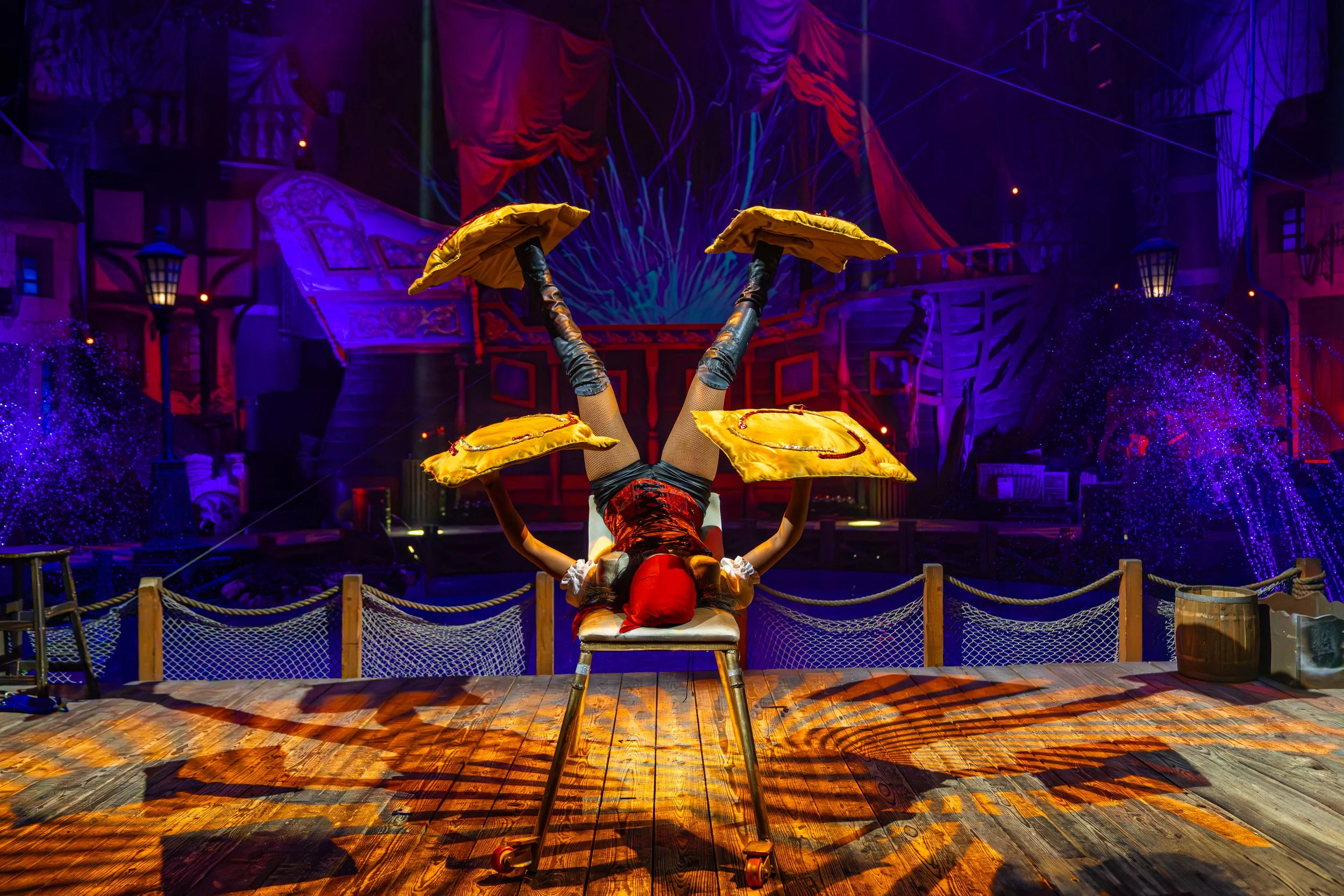 A person performing a daring upside-down balancing act on a chair on a theatrical stage, with colorful props and scenery in the background.