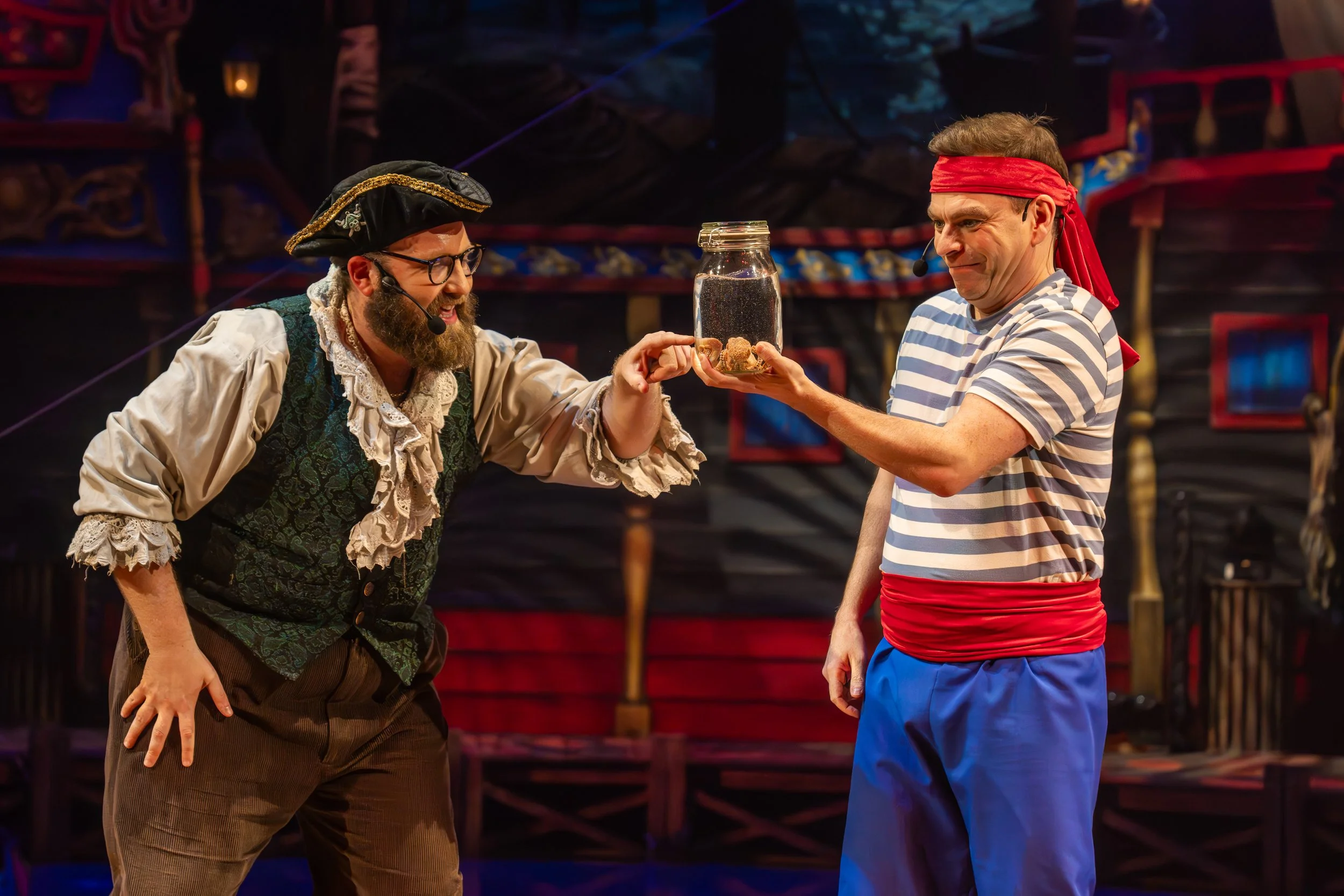 Two actors on stage in costume, one dressed as a pirate and the other as a sailor, sharing a humorous moment with a jar of what appears to be dirt or rocks.