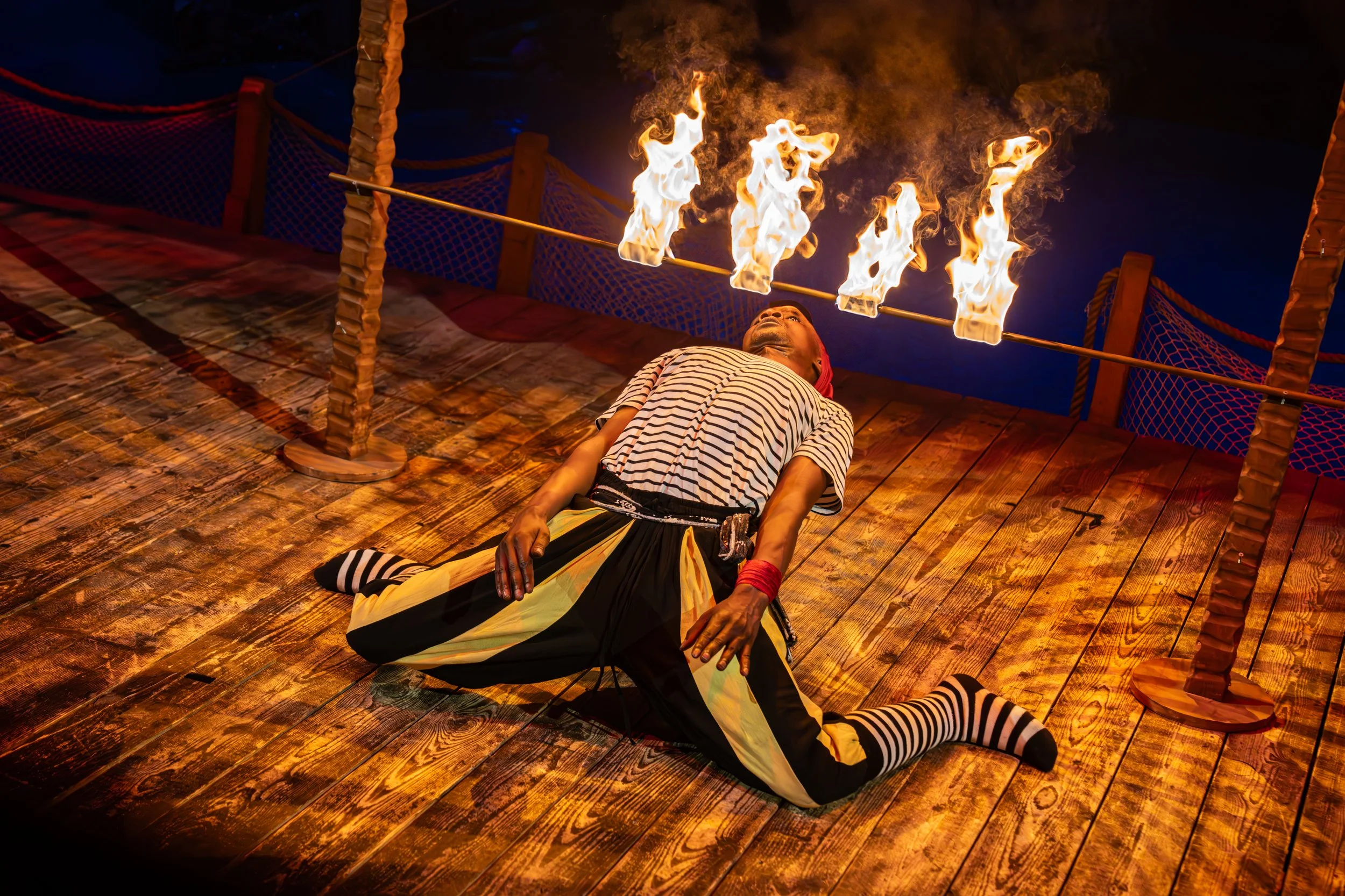 Performer lying on wooden stage with flames on a horizontal pole overhead during a fire show or act.