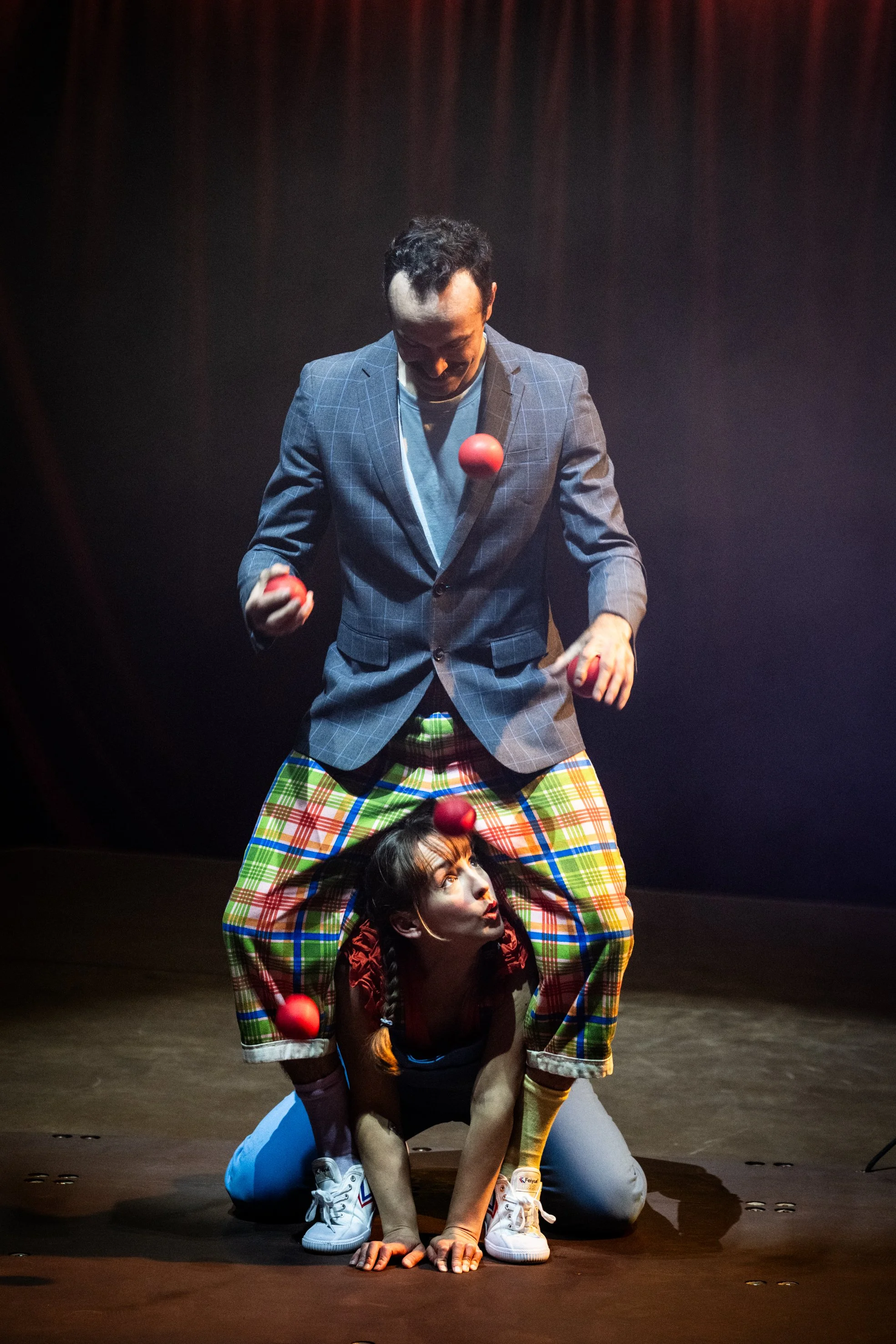 Two performers, one standing and one kneeling, juggling red balls on stage with a dark background.