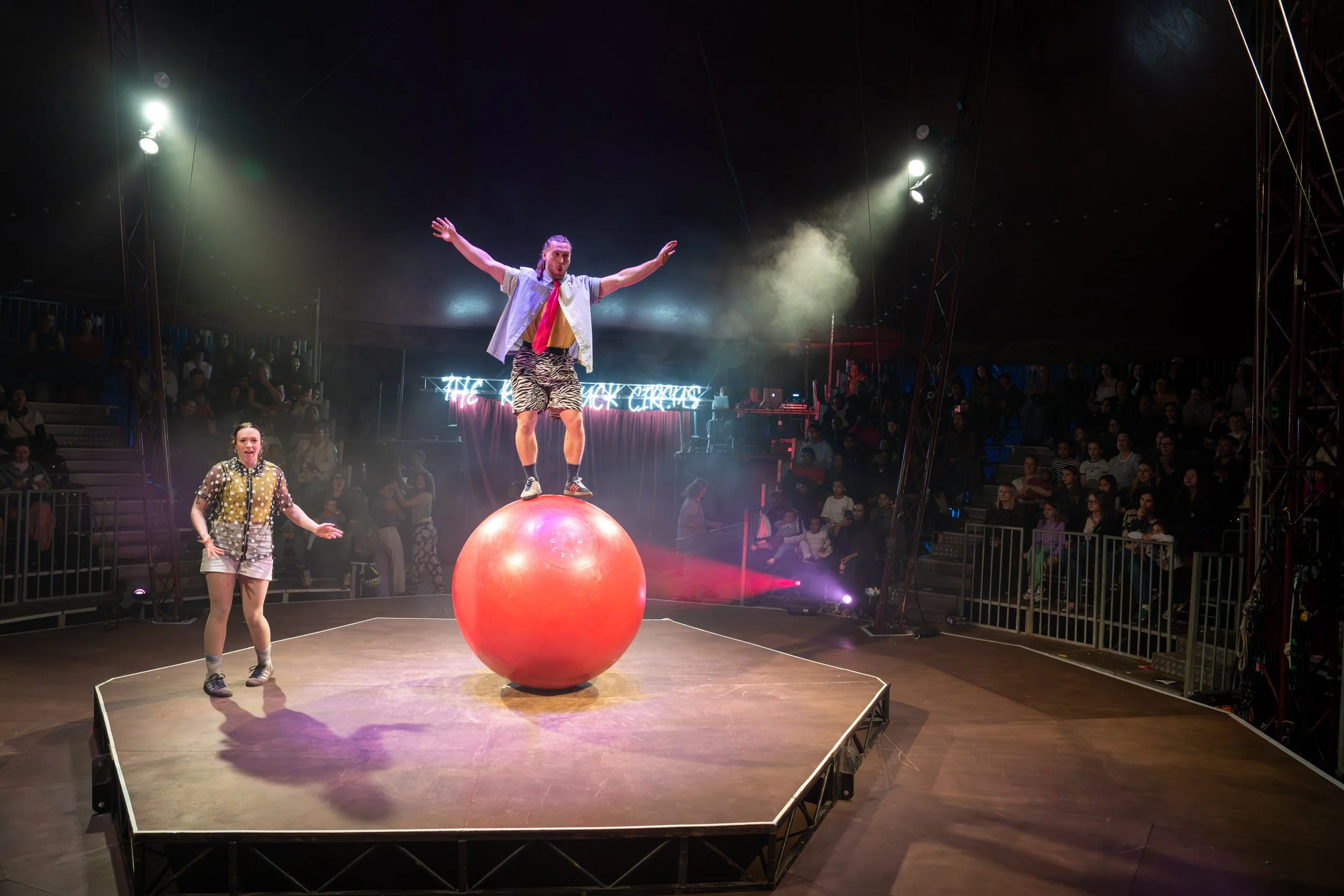 Circus performers balancing on a large red ball under bright stage lights while an audience watches from the background.