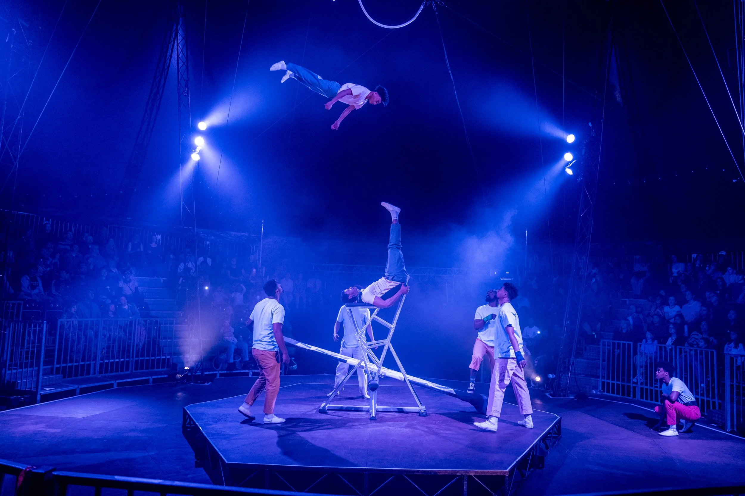 Circus performers perform acrobatics on a stage with blue lighting, including a man in a white shirt holding another performer upside down, while others watch and the audience observes from the background.