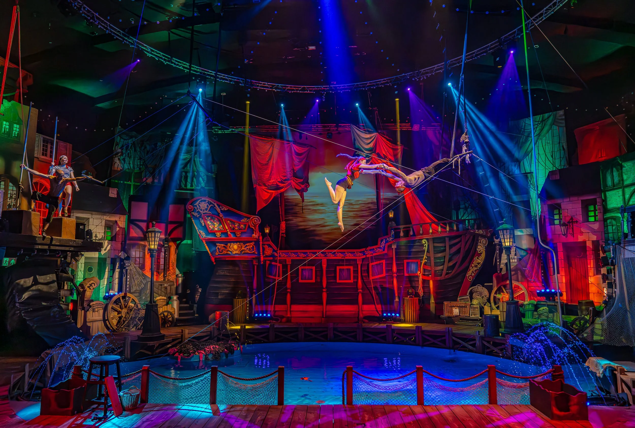 Indoor circus stage with performers trapeze artist and acrobats, colorful lights, and a set resembling a pirate ship.