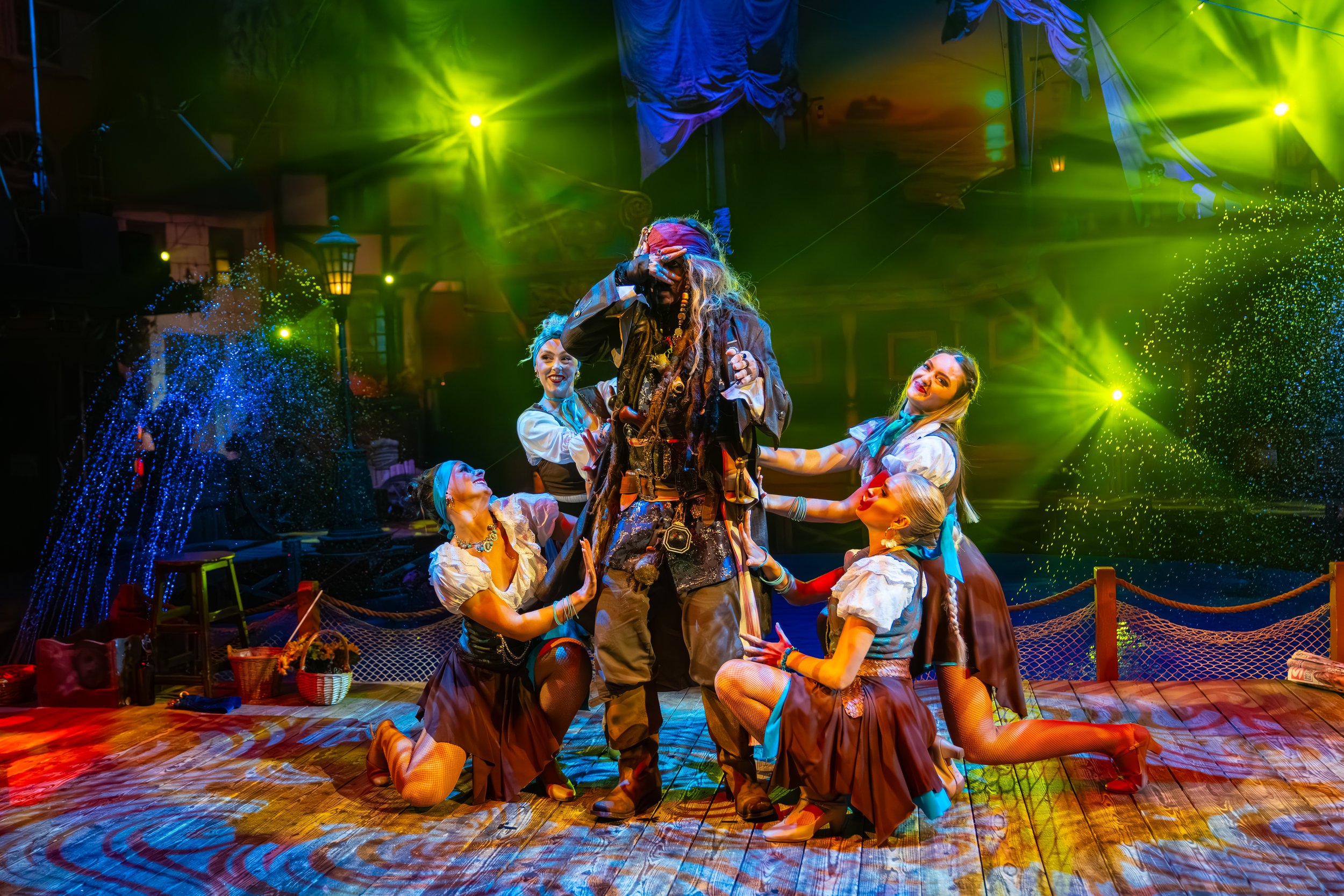 A theatrical stage scene featuring a pirate character with dreadlocks, surrounded by five women dressed in pirate-themed costumes, with colorful stage lights and water spray effects in the background.