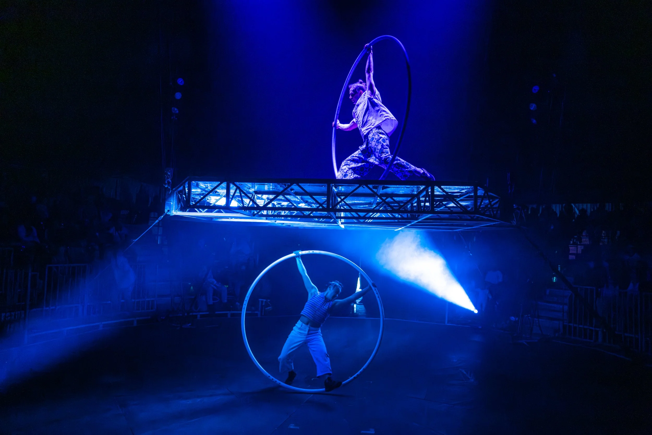 Two acrobats performing circus acts with hoops, illuminated by blue lighting in a dark arena with an audience.