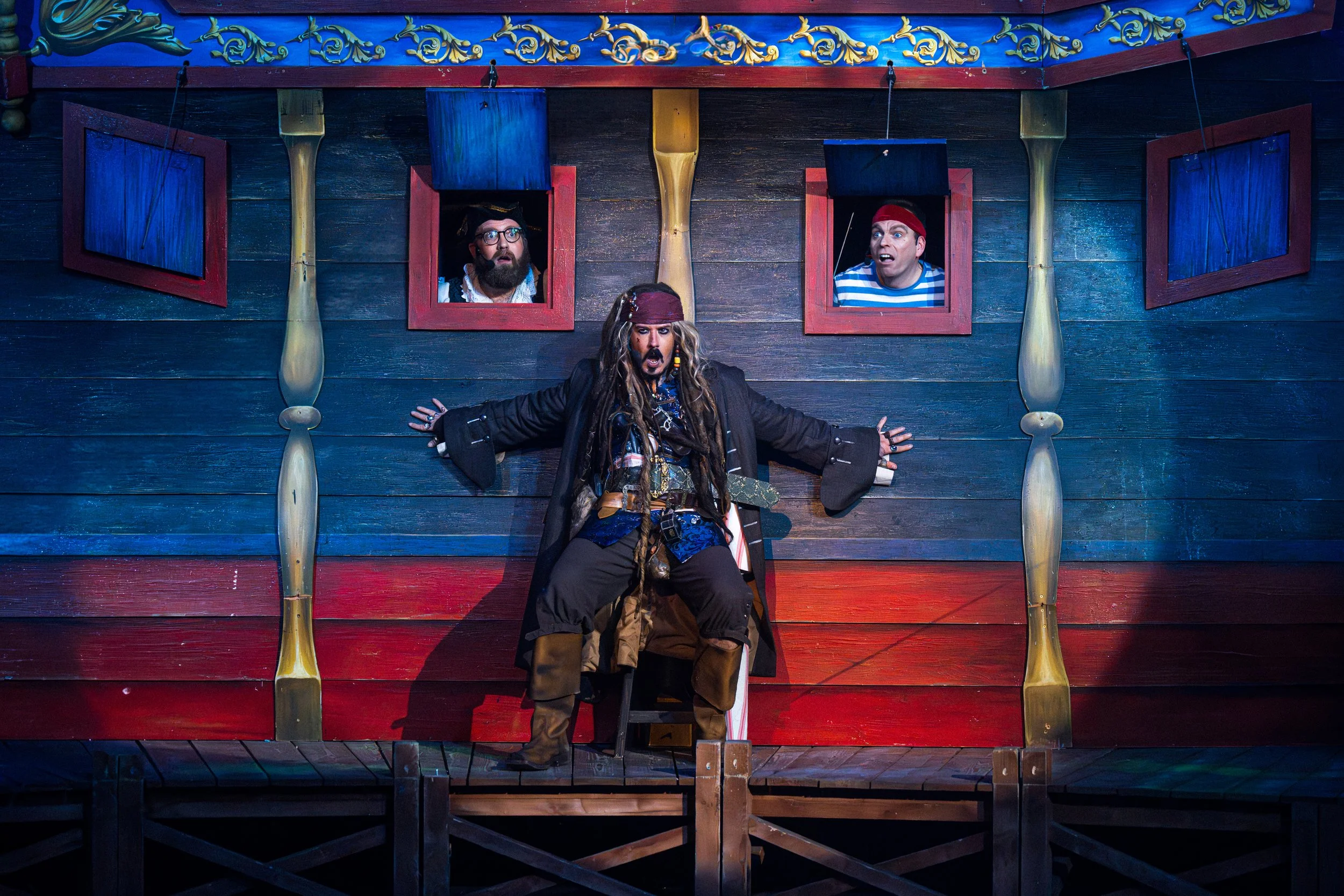 A theatrical performance with a pirate actor on stage dressed in pirate costume, seated with arms outstretched, in front of a wooden backdrop with three windows. Two actors look out from the windows with surprised expressions.