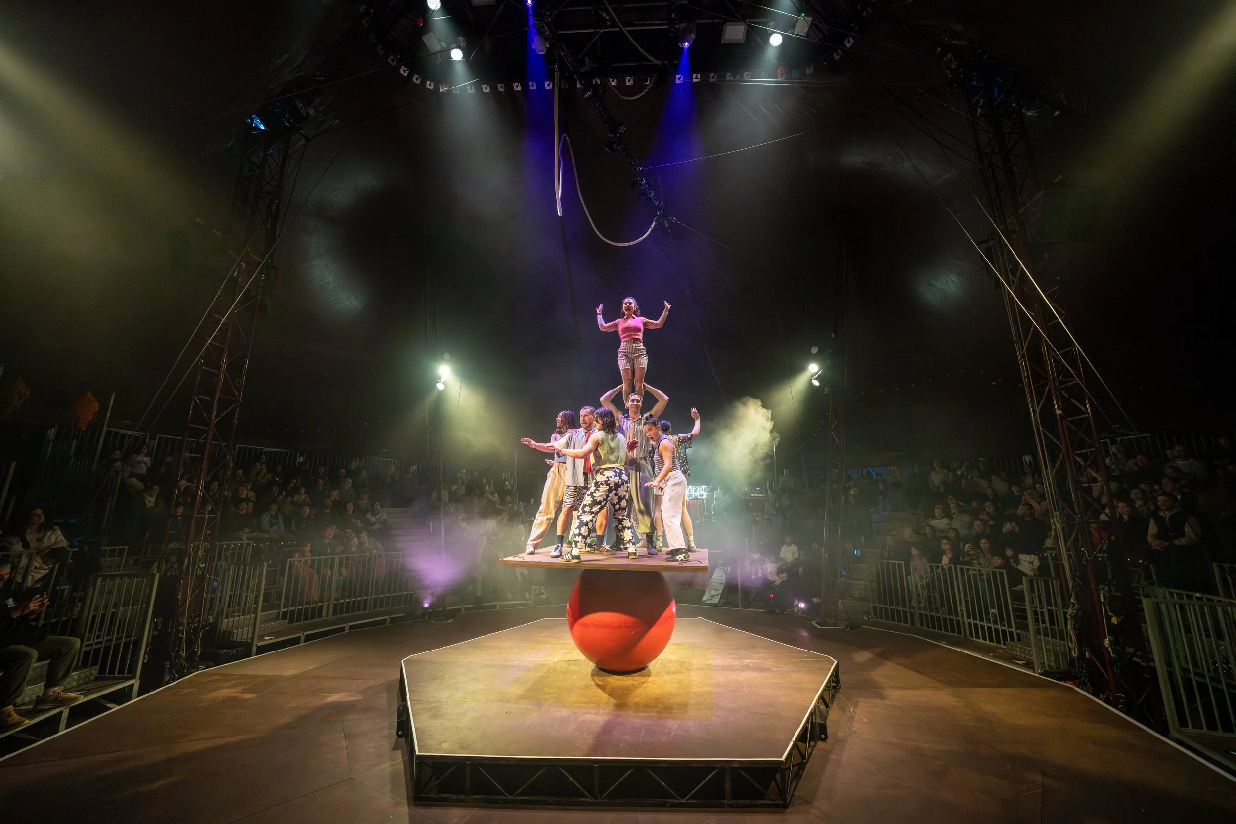 A group of performers in colorful costumes forming a human pyramid on a stage in an indoor circus or performance venue, with an audience watching from the sides and stage lighting illuminating the scene.