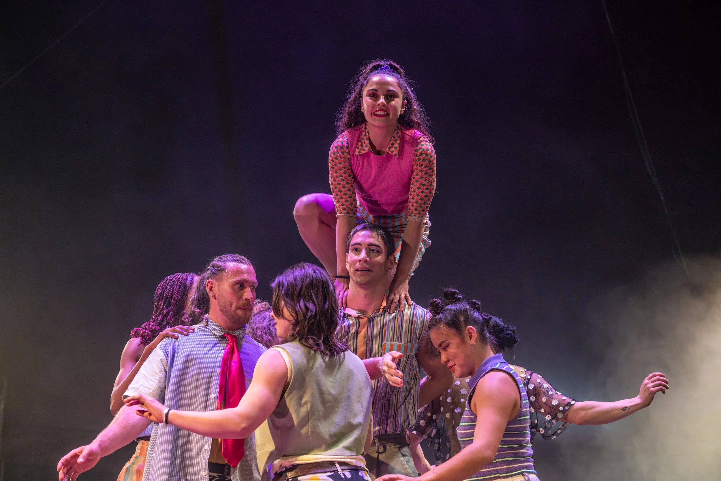 Six performers on stage, five standing and one crouching on their shoulders, with purple and yellow stage lighting.