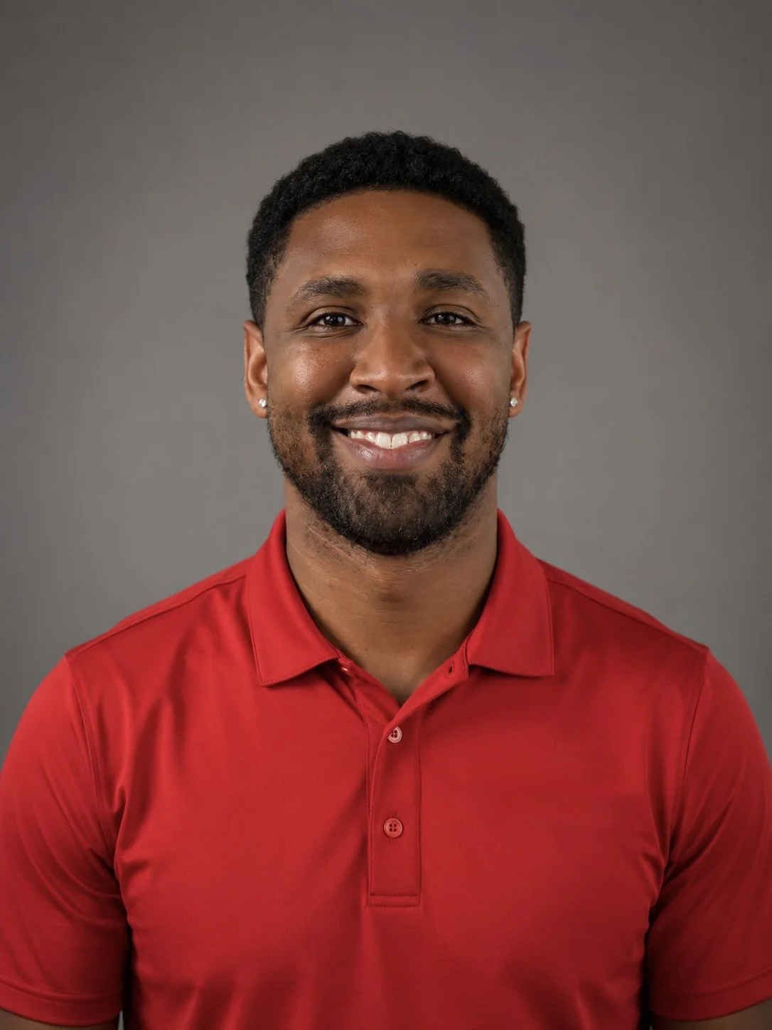 A smiling man with a beard and short curly hair, wearing a red polo shirt, against a gray background.