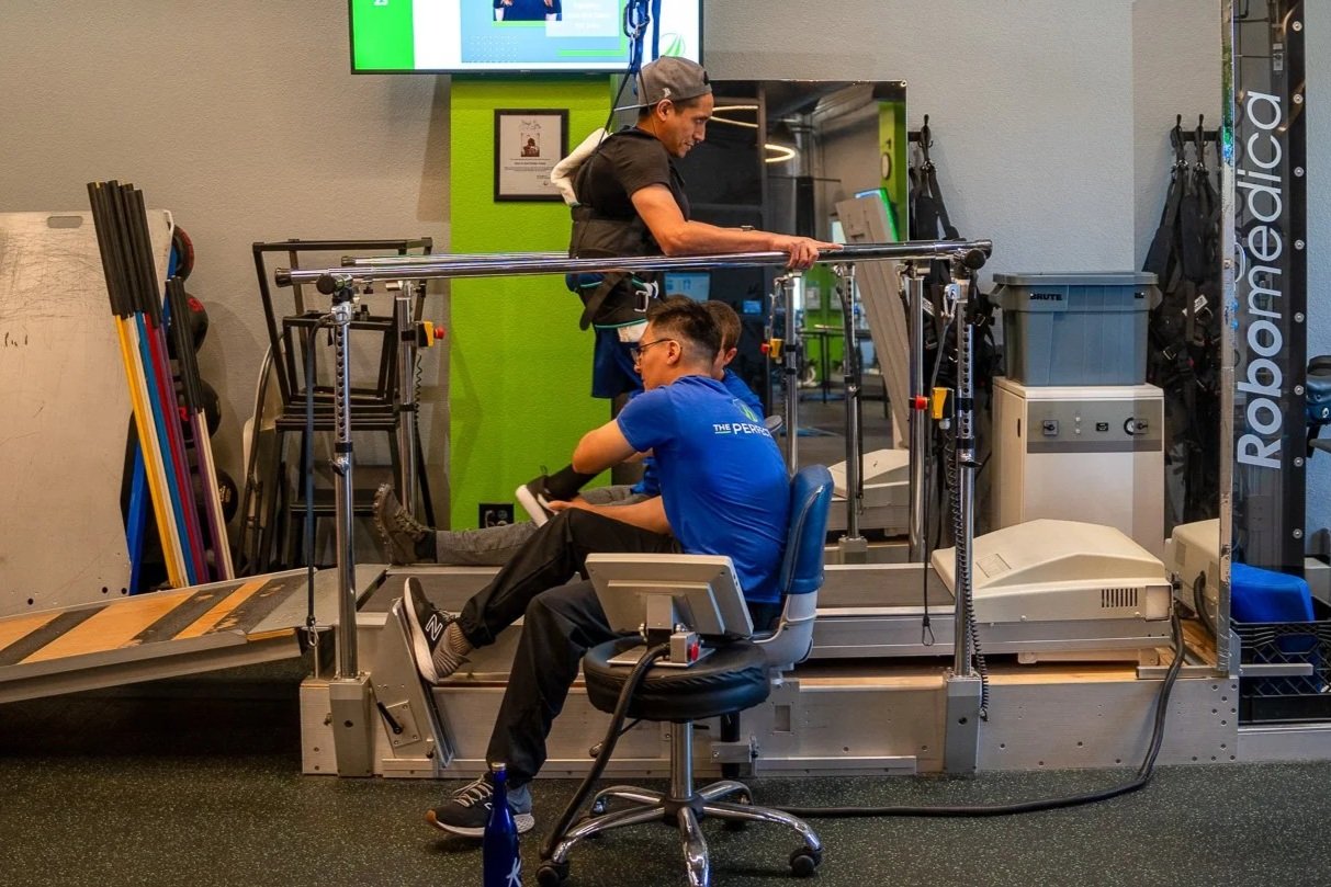 A person in a black shirt and gray cap assists a man in a blue shirt with a leg injury on a specialized rehabilitation treadmill with a harness system in a gym. The physician is standing and the man is seated on a chair with a computer monitor nearby.