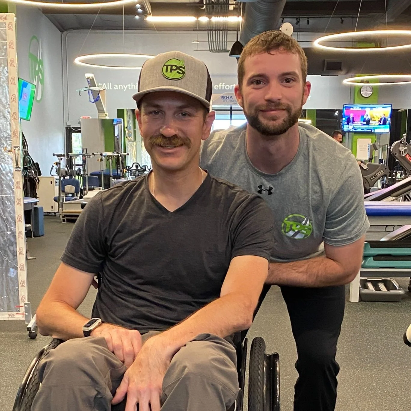 Two men posing for a photo inside a gym, one in a wheelchair in a black t-shirt and gray cap, the other standing behind him in a gray t-shirt with the gym logo.