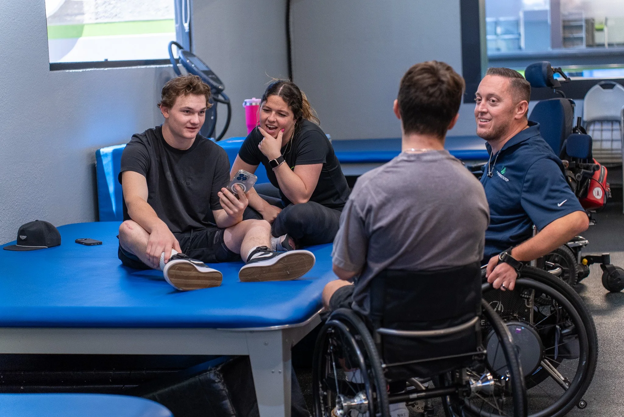 Four people, two young men and two young women, are sitting on a blue mat in a gym or physical therapy center. They are engaged in conversation and smiling, with one young man holding a smartphone. The group includes a young man in a wheelchair and a man in a motorized wheelchair, both wearing dark gray and navy-blue shirts respectively. The setting has large windows and exercise equipment in the background.