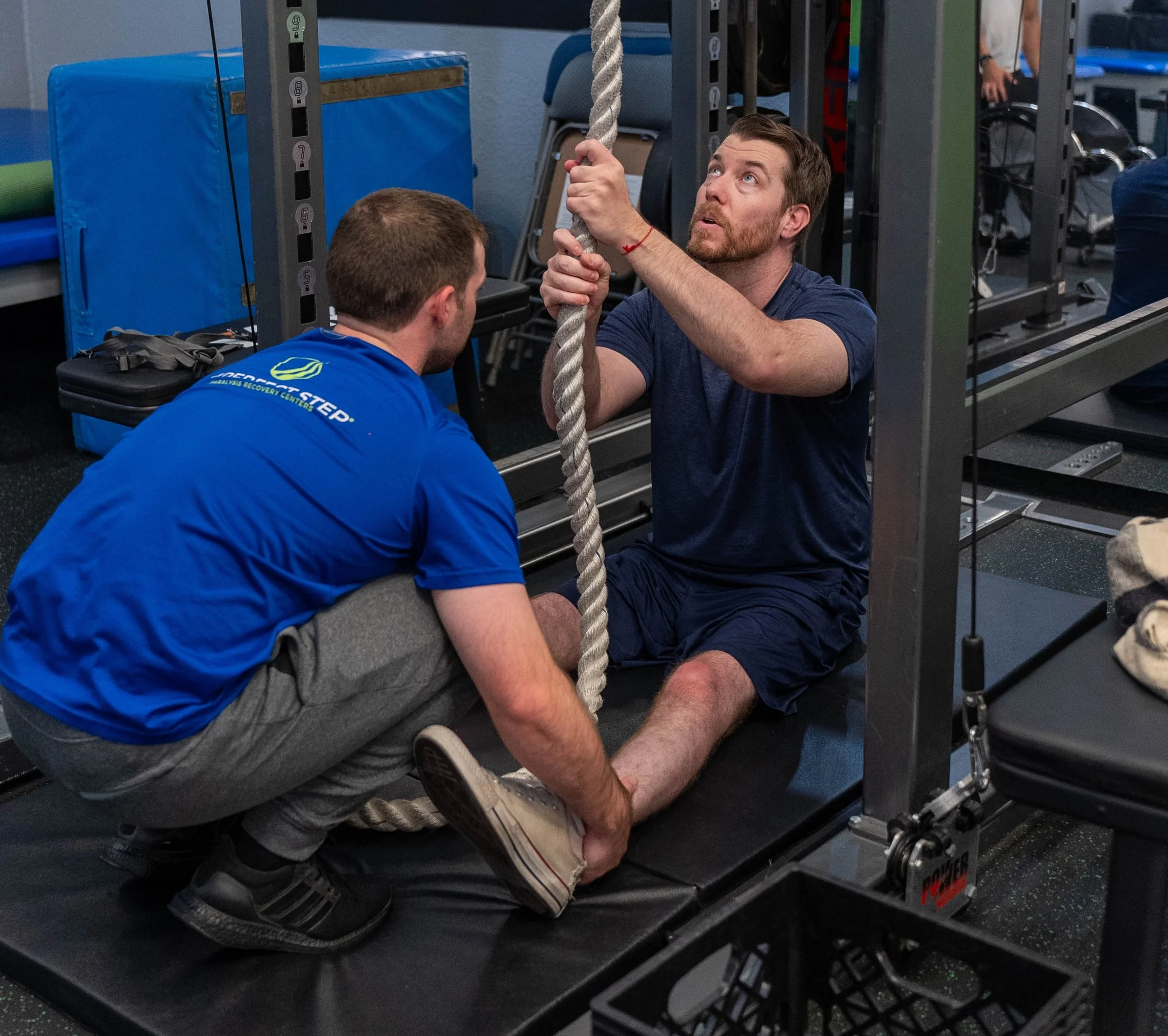 A man receiving physical therapy or assistance with a rope in a gym, while another person provides support.