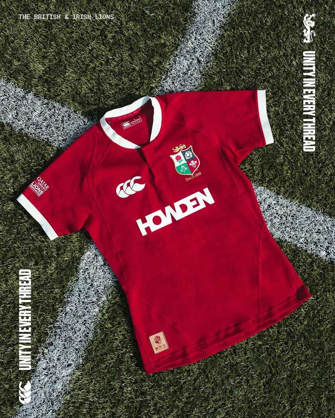 Red rugby jersey with white collar and sleeve trim, placed on a grassy field with white lines. The jersey features a crest with a shield, a crown, and floral emblem, a logo with a stylized 'C', and the word 'HOWDEN' in white across the front. There i