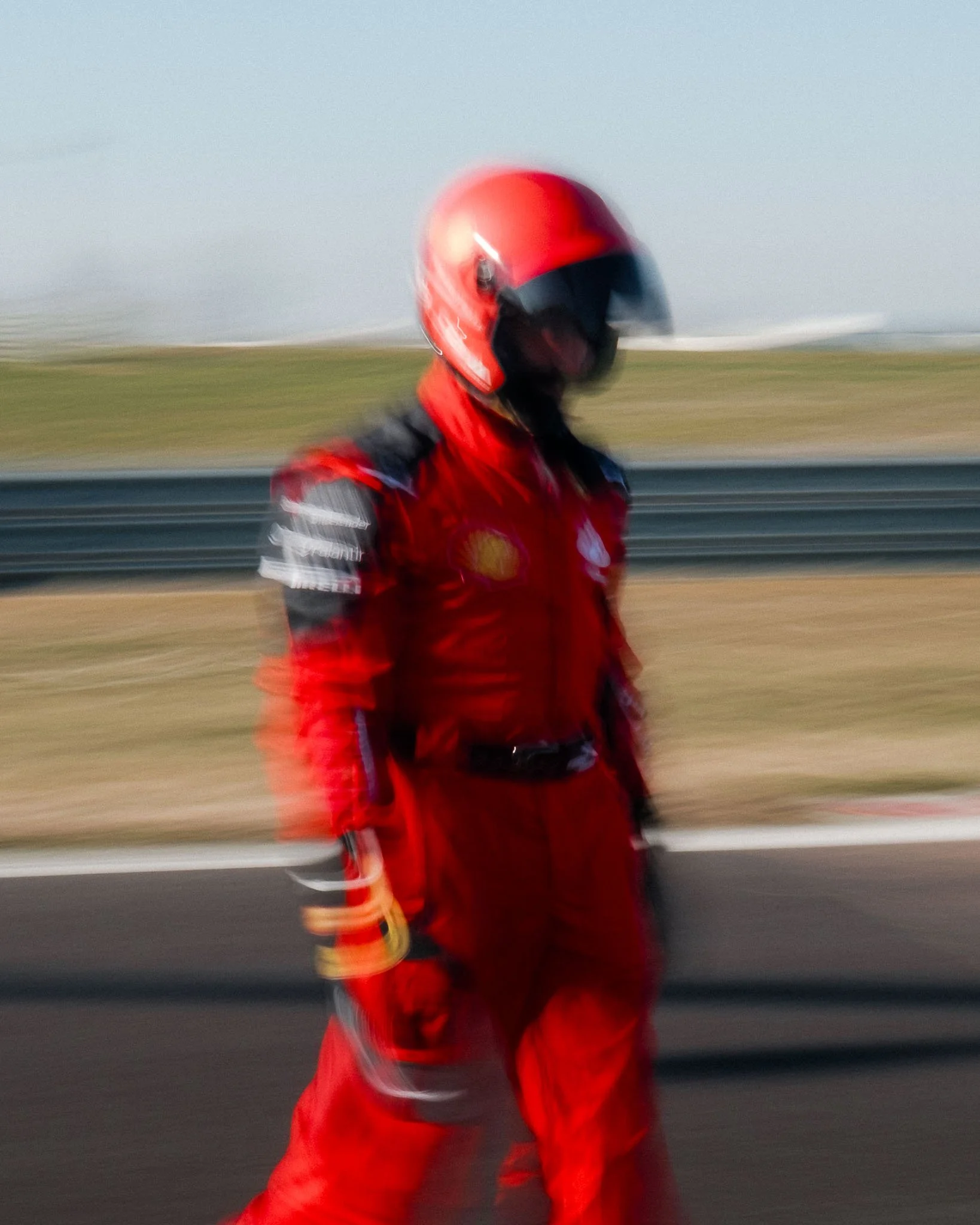 A person in red racing gear walking on a racetrack, wearing a red helmet and a red racing suit with various sponsor patches.